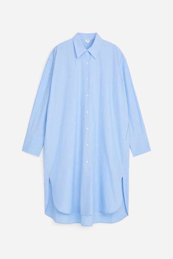 Une robe-chemise bleu clair à coupe ample est présentée de face, arborant un motif rayé subtil, un col classique, une fermeture boutonnée sur toute la longueur, et des manches longues avec poignets boutonnés. La robe mi-longue est conçue avec un ourlet arrondi, des fentes latérales, et un panneau arrière légèrement plus long que l'avant. - 1
