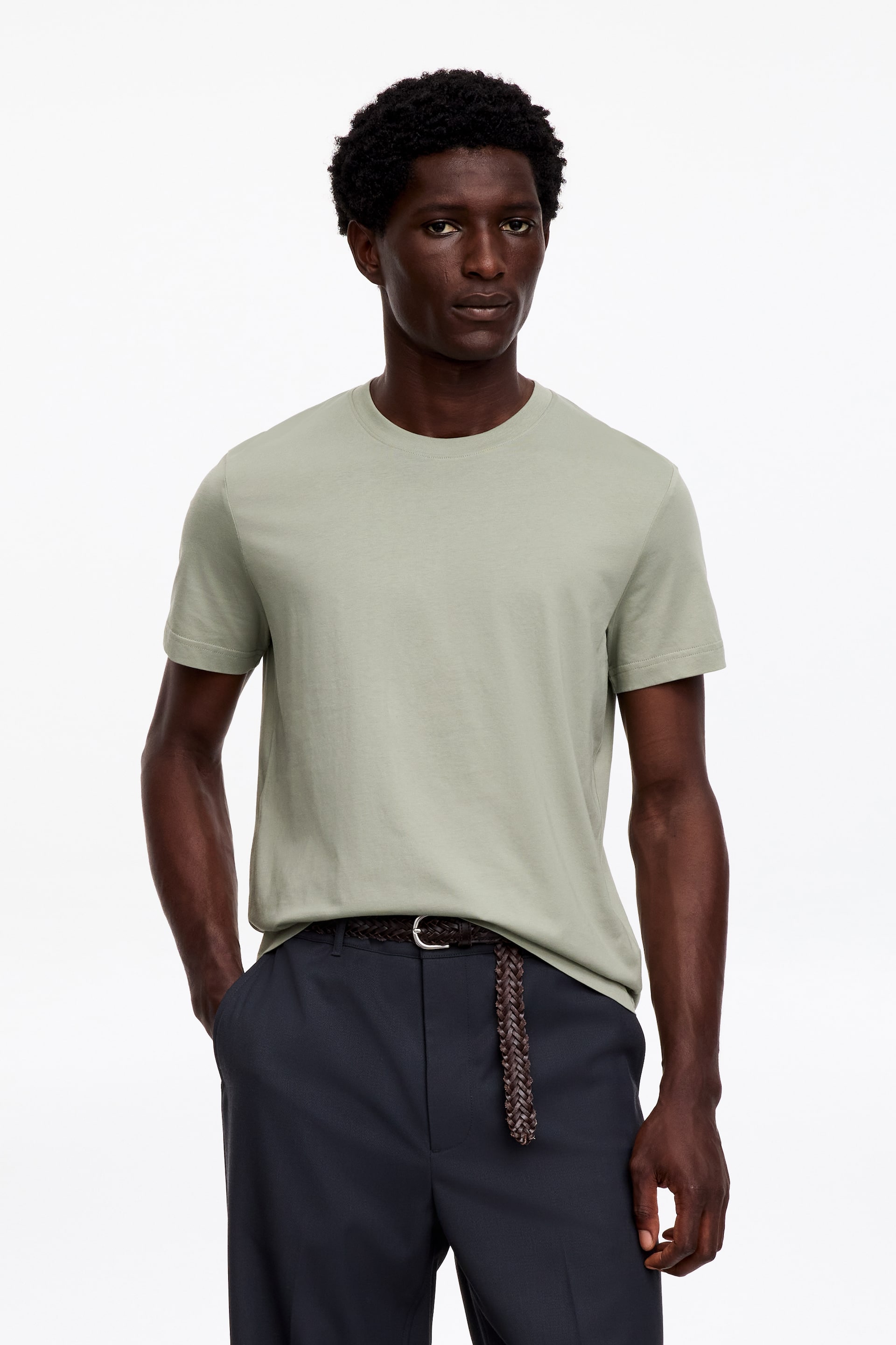 Grotere afbeelding bekijken: Een lichtgroen T-shirt met ronde hals wordt gedragen door het model, met een getailleerde pasvorm en iets kortere, aansluitende mouwen en een gladde textuur. Het T-shirt is ingestopt in een donkere broek, vastgezet met een gevlochten bruine riem.