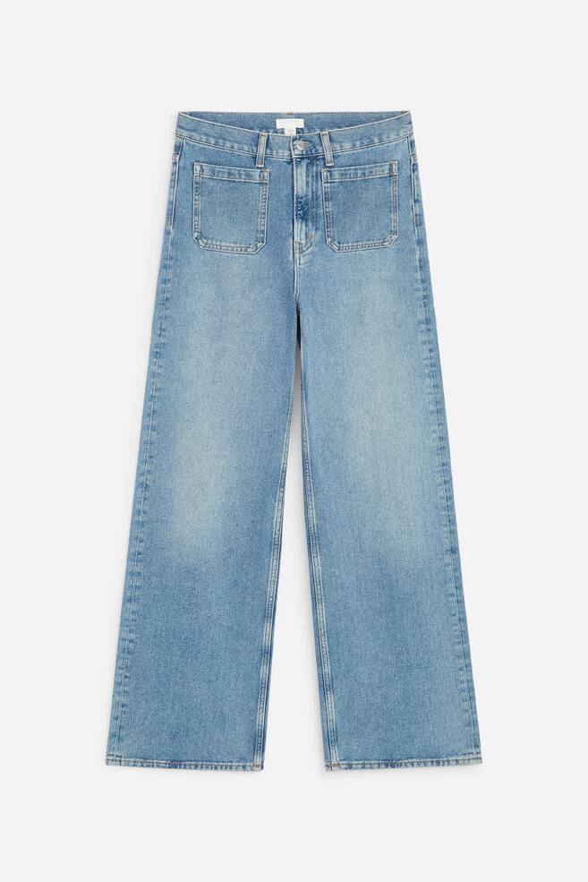 A pair of blue denim jeans is presented flat, facing FRONT, featuring a high-rise waist and a zip fly. It is designed with a slim fit through the upper leg, flaring into wide legs, and includes large rectangular patch pockets on the front. - 1