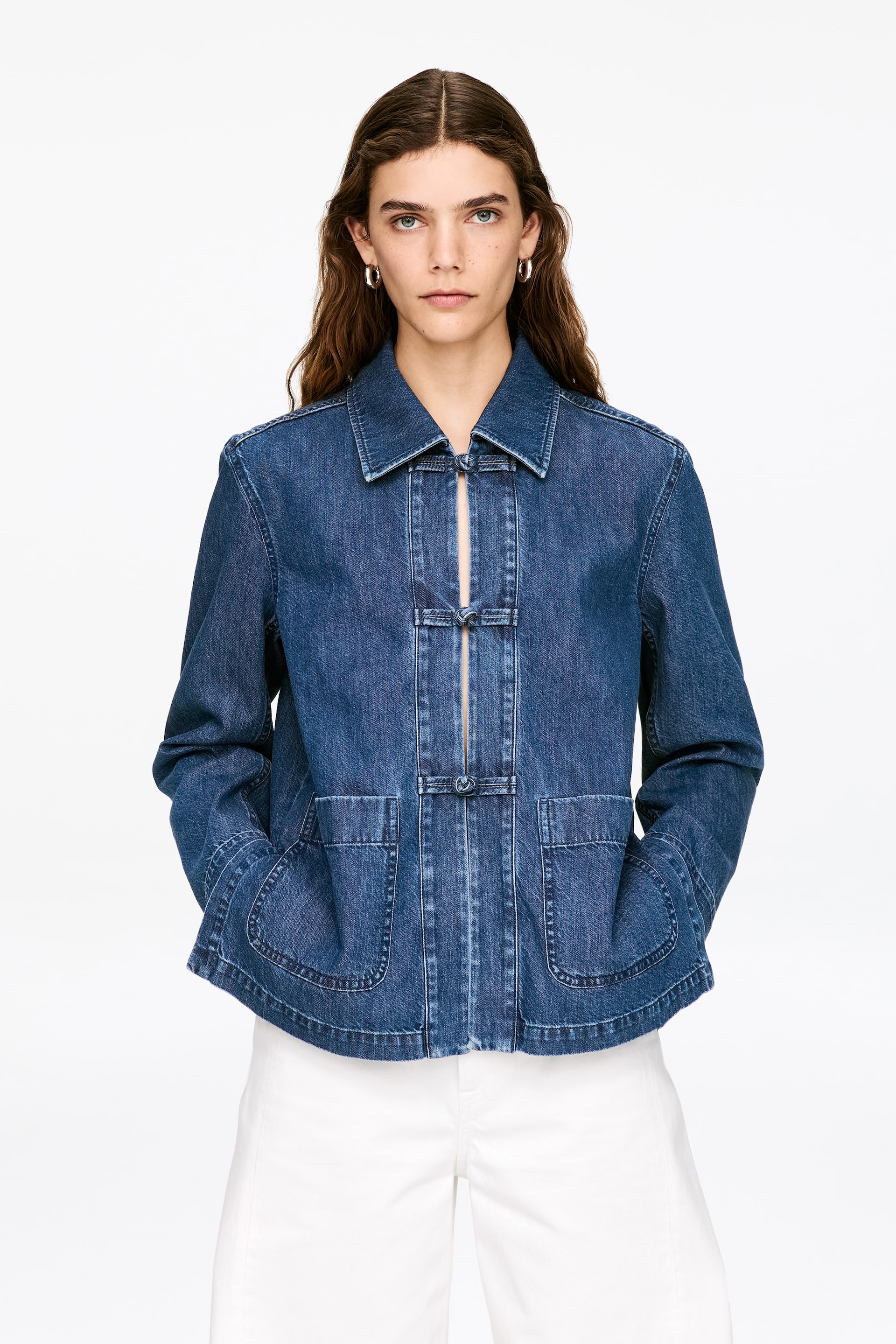 View larger image: A denim jacket featuring a pointed collar and three central frog closures down the front. It also displays two large, rounded patch pockets at the waist, presented front-facing.