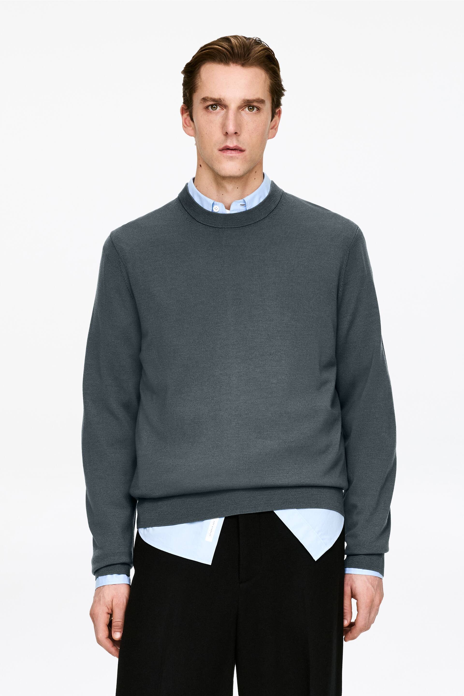 Agrandir l'image: Un pull à col rond gris foncé est présenté de face, mettant en avant une texture en maille lisse et des poignets et ourlets côtelés. Il est superposé sur une chemise à col bleu clair, visible au niveau du col, des poignets et de l'ourlet inférieur.