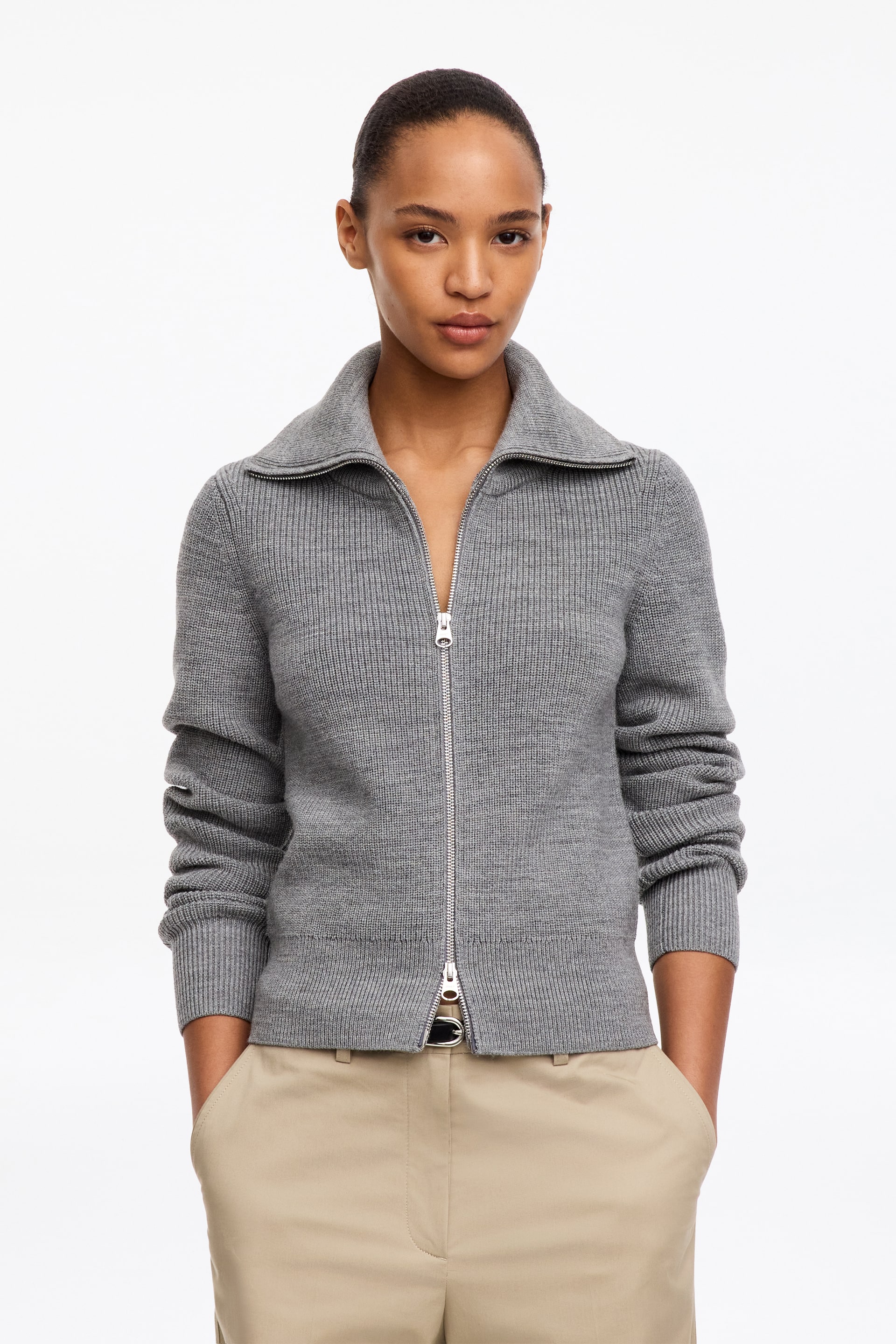 Grotere afbeelding bekijken: Het grijze Wool-Cotton Zip Cardigan, met een ribgebreide structuur en een brede kraag, wordt gedragen door het model. De zilverkleurige rits is aan beide uiteinden gedeeltelijk open geritst, en het cardigan is gestyled over een lichtgekleurde broek.
