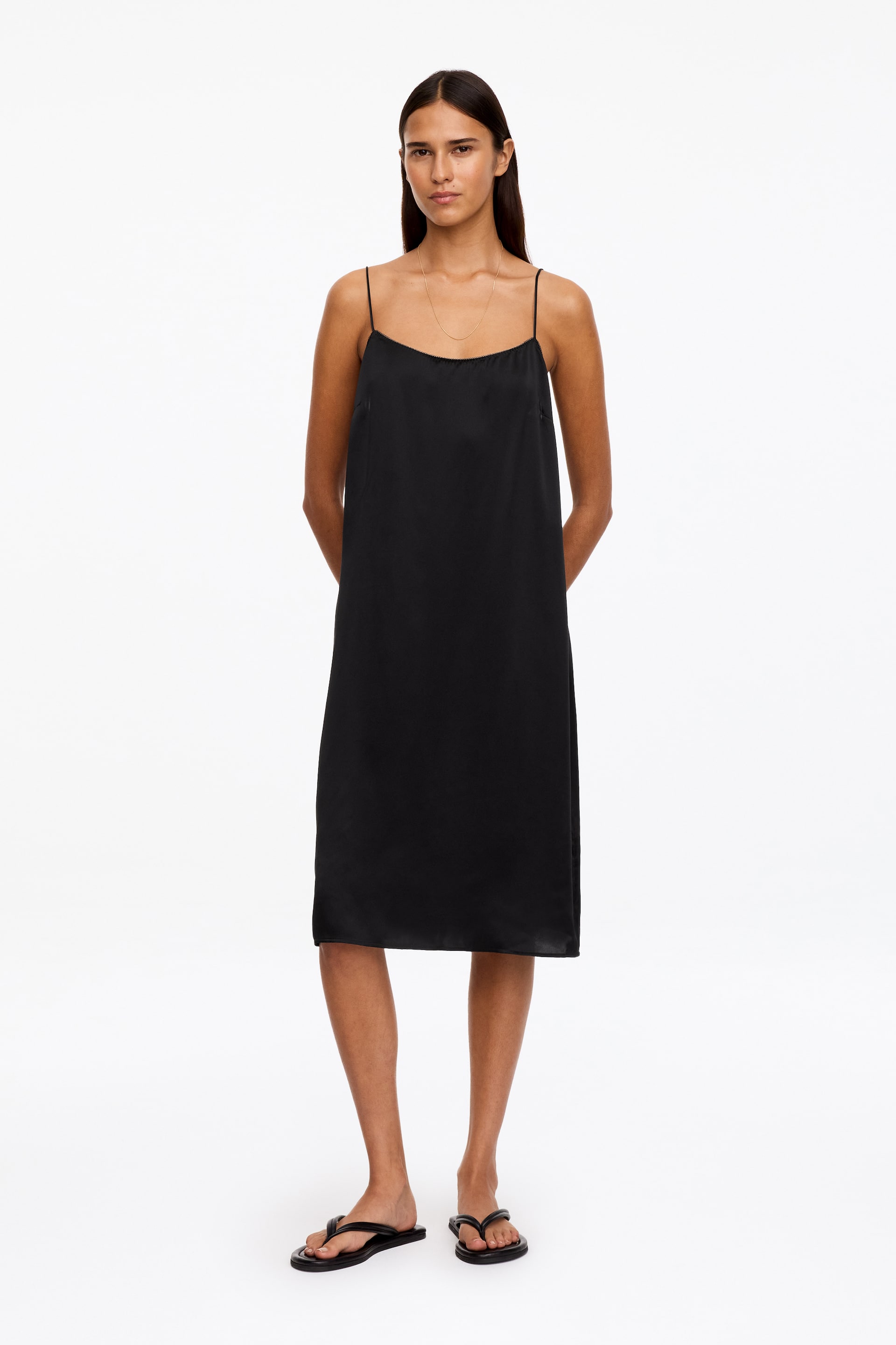 View larger image: A black midi slip dress with a straight neckline, delicate shoulder straps and a relaxed silhouette is being worn by the model. The dress falls below the knee and is styled with black flat thong sandals.