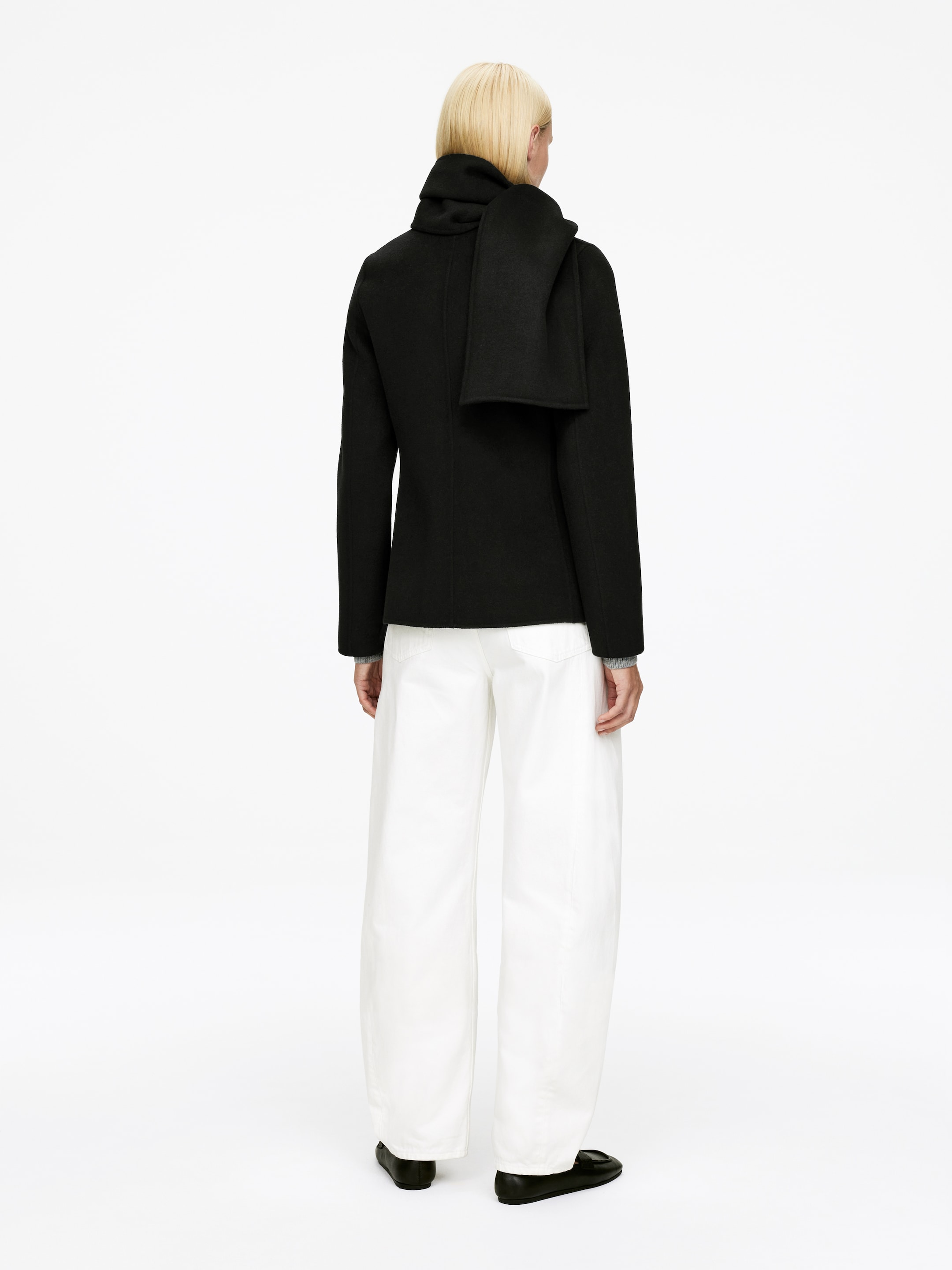 Agrandir l'image: Une veste courte noire avec un col haut est vue de dos, tandis qu'une écharpe noire est drapée autour du cou avec une extrémité tombant dans le dos. La veste est portée avec un pantalon blanc à jambes larges et des chaussures plates noires.