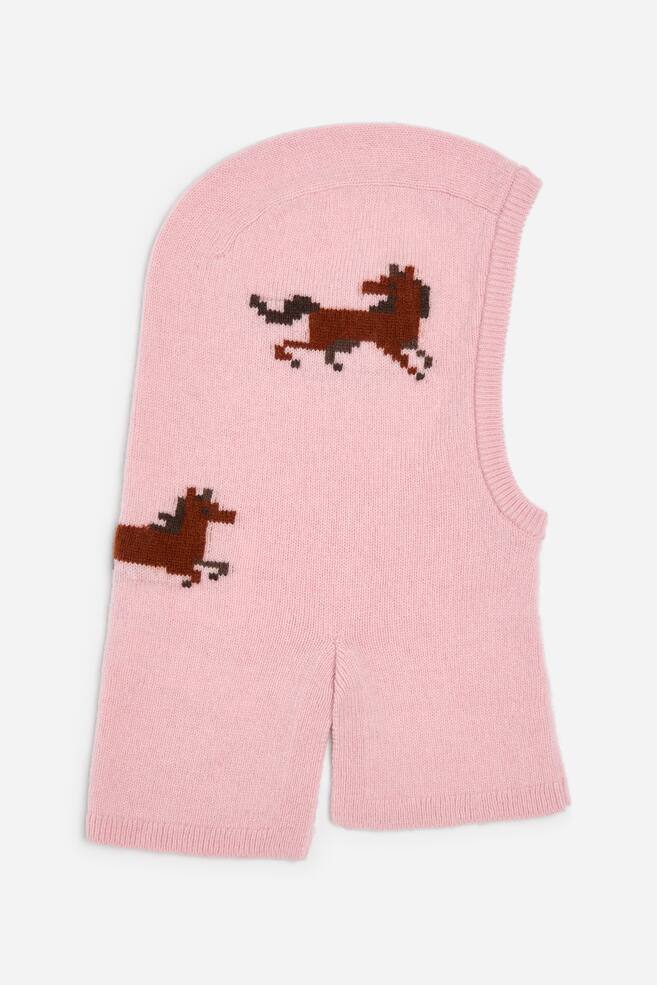 A light pink knitted balaclava hood is shown from the side, featuring two brown pixelated horse graphics knitted into the fabric. The hood is designed with a ribbed trim around the face opening and a split hem at the bottom. - 1
