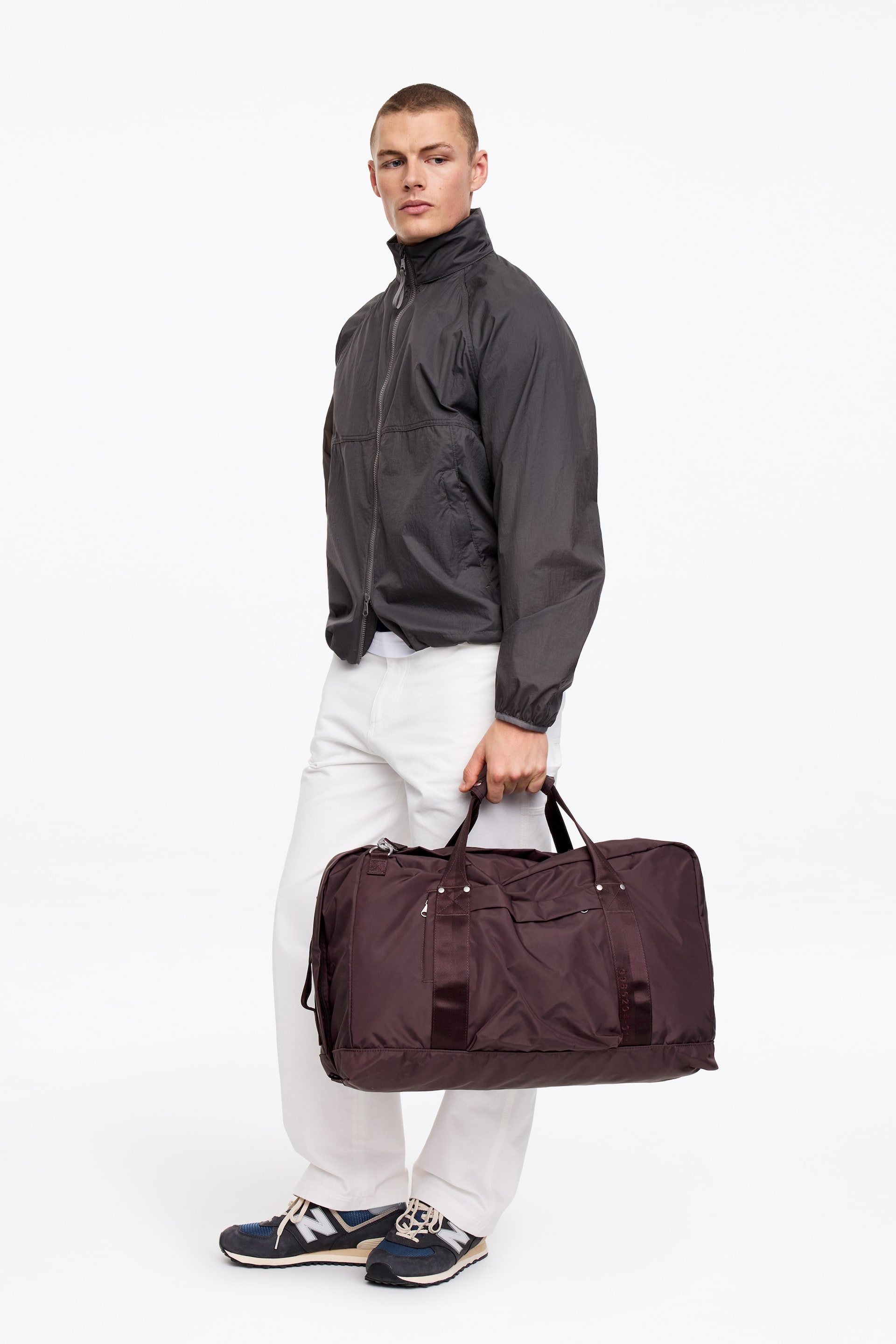 Visa större bild: En mörkbrun nylonväska bärs i sina två övre handtag, något framåtlutad och åt höger. Den har en rektangulär form med en stor främre ficka och ett vertikalt banddetalj på framsidan, tillsammans med synlig silverfärgad hårdvara.