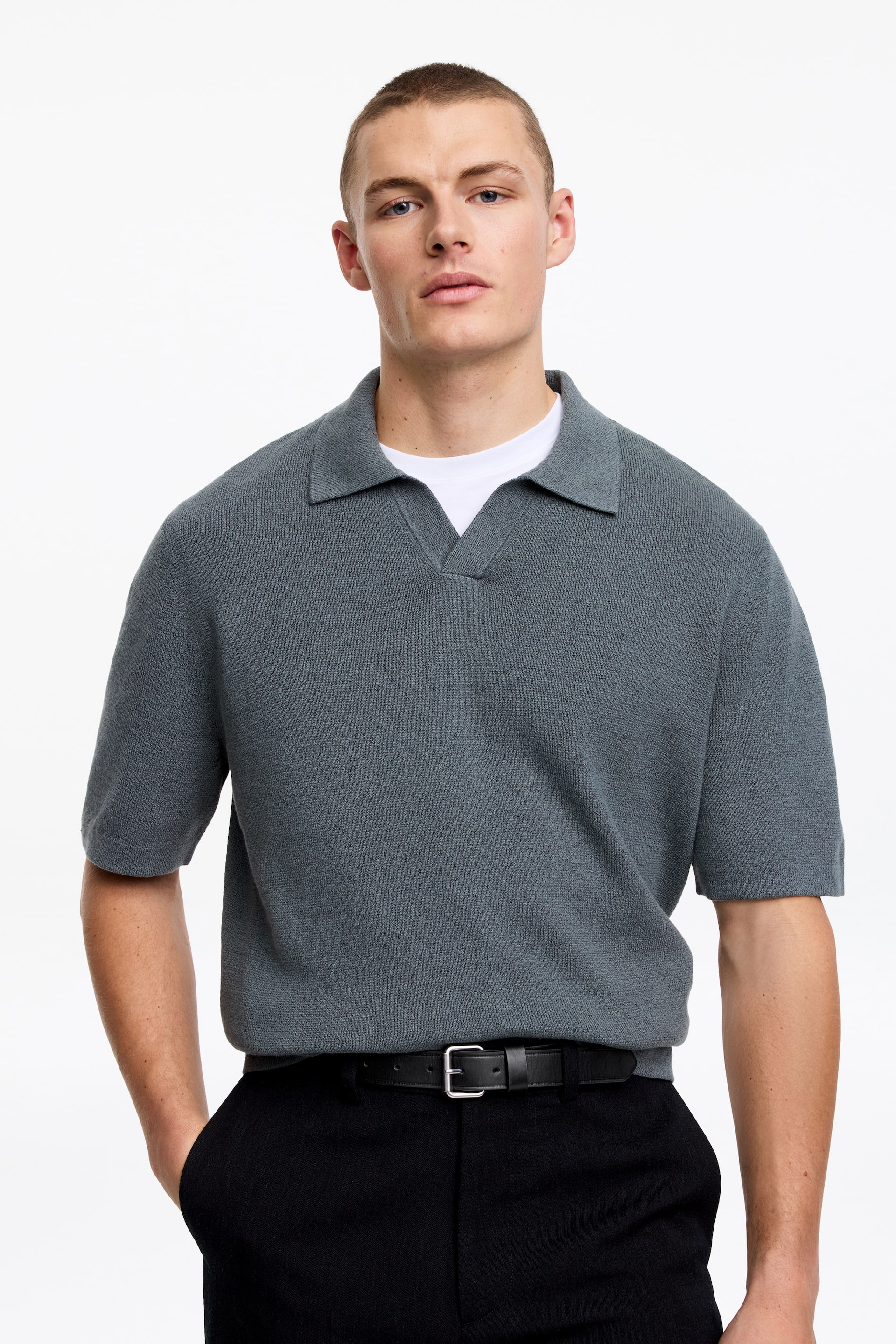 View larger image: A grey knitted polo shirt is being showcased, featuring short sleeves and a spread collar with an open V-neck placket. A white T-shirt is visibly layered underneath the polo.