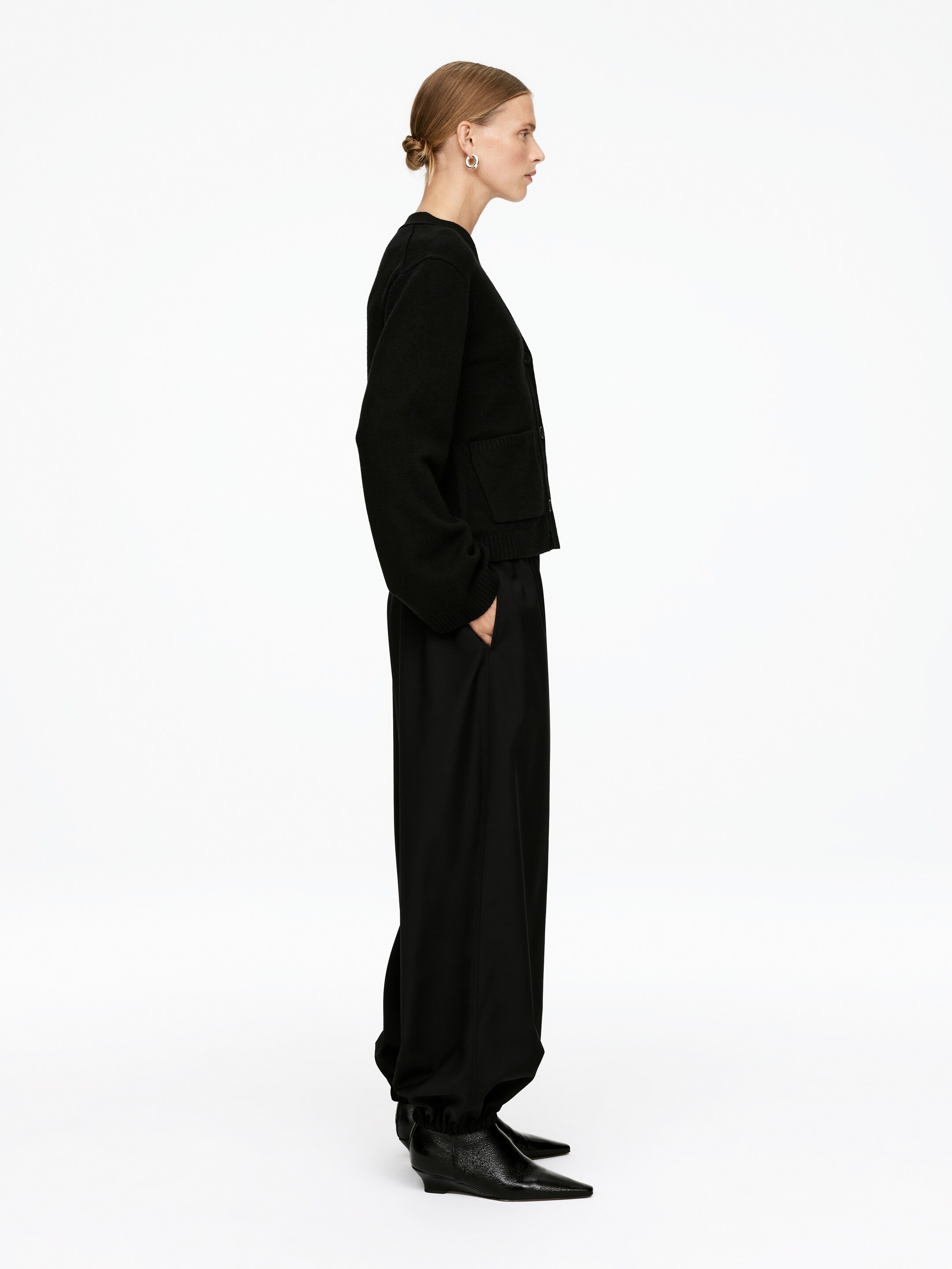View larger image: A black knit cardigan and wide-leg trousers are being presented in a side profile. The cropped cardigan is featuring long, full sleeves with two visible patch pockets and a ribbed hem, while the loose-fitting trousers are gathering at the ankles, creating a balloon effect over pointed-toe boots.