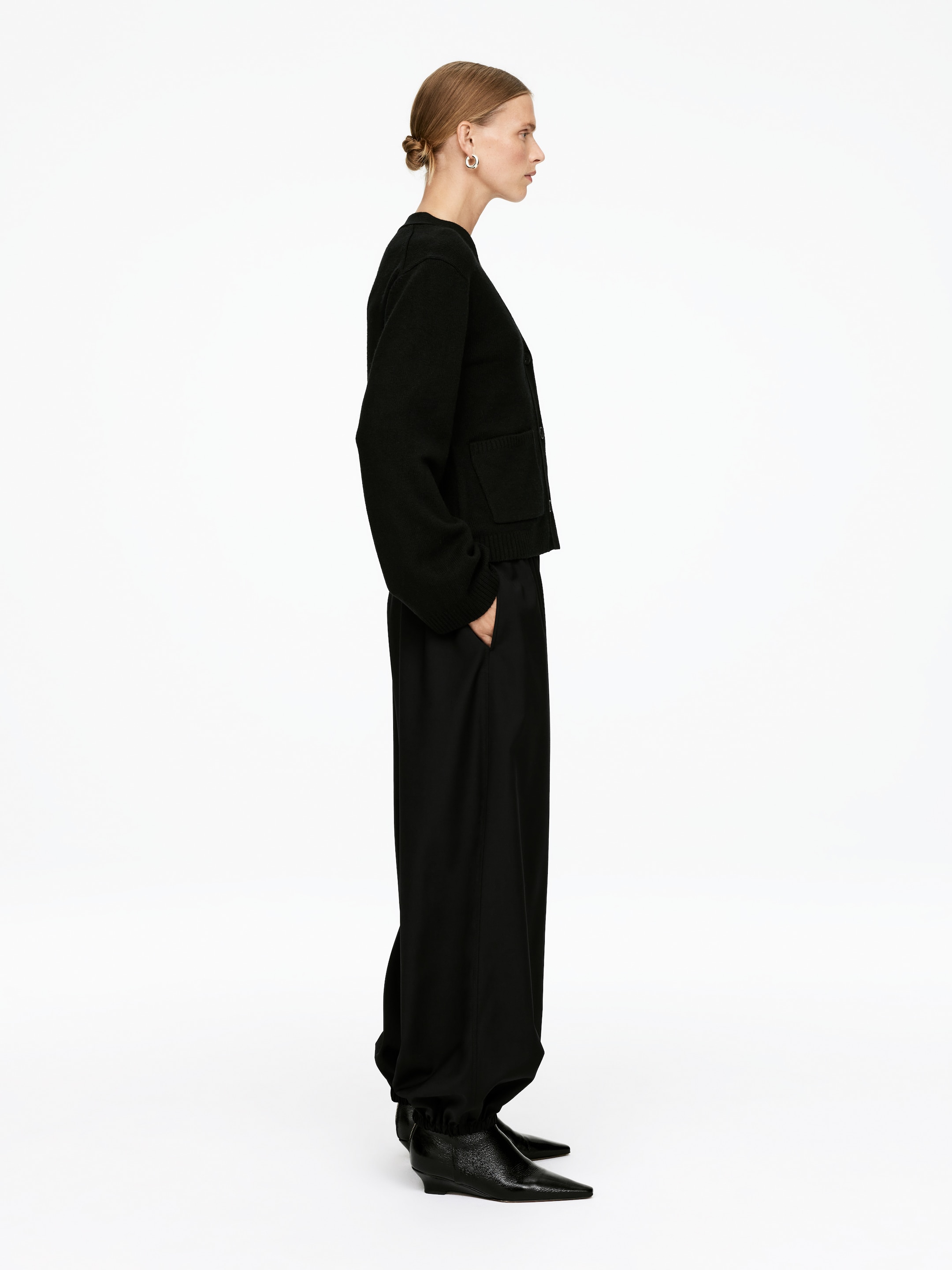 Größeres Bild anzeigen: Eine schwarze Strickjacke und weite Hosen werden im Seitenprofil präsentiert. Die kurze Strickjacke hat lange, volle Ärmel mit zwei sichtbaren aufgesetzten Taschen und einen gerippten Saum, während die locker sitzenden Hosen an den Knöcheln gerafft sind und einen Balloneffekt über spitzen Stiefeln erzeugen.