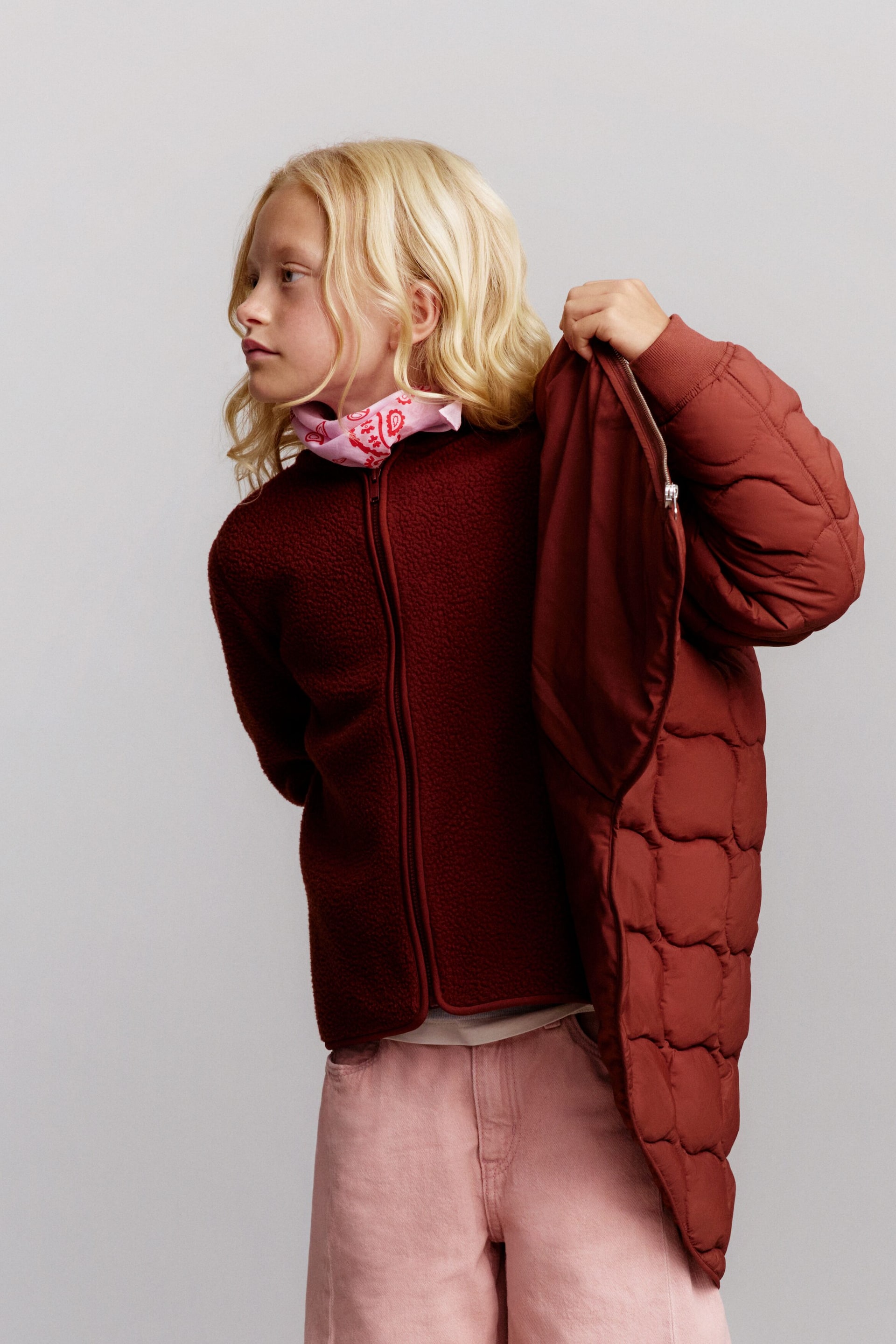 Visa större bild: En mörkt rödbrun fleecejacka med ståkrage och jerseykantning bärs i en trekvarts frontvinkel, kombinerad med ljusrosa barrel-jeans och en rosa paisley-bandana. En matchande roströdbrun quiltad lättviktsjacka draperas över axeln och lyfter fram dess vågformade quiltade design.