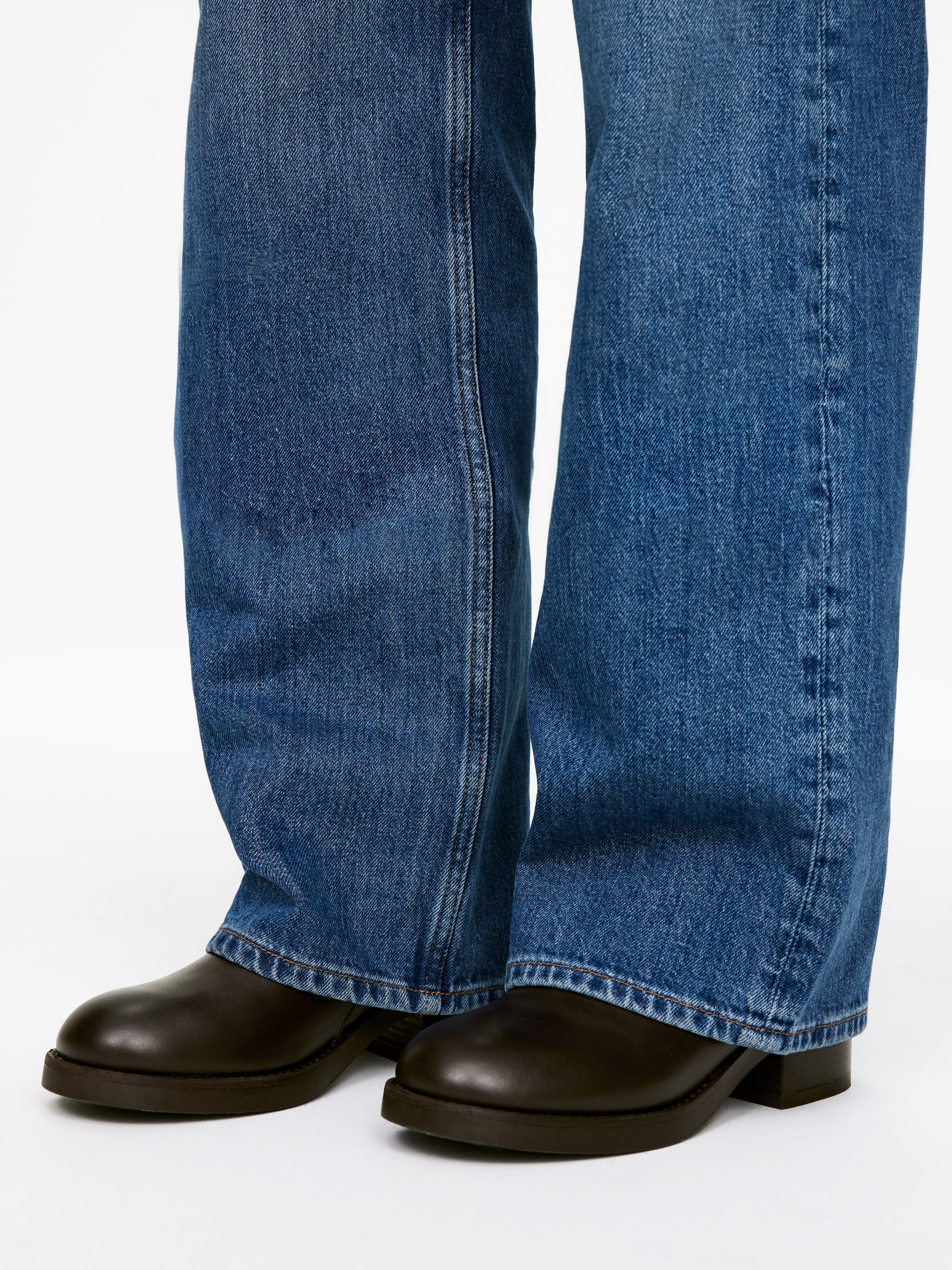 Agrandir l'image: Un jean en denim bleu tombe sur des chaussures en cuir marron foncé. Le jean présente une silhouette à jambes larges, et des coutures visibles descendent le long de l'avant de chaque jambe. Les chaussures, qui sont légèrement orientées vers l'extérieur, présentent un bout arrondi et un talon bas empilé.