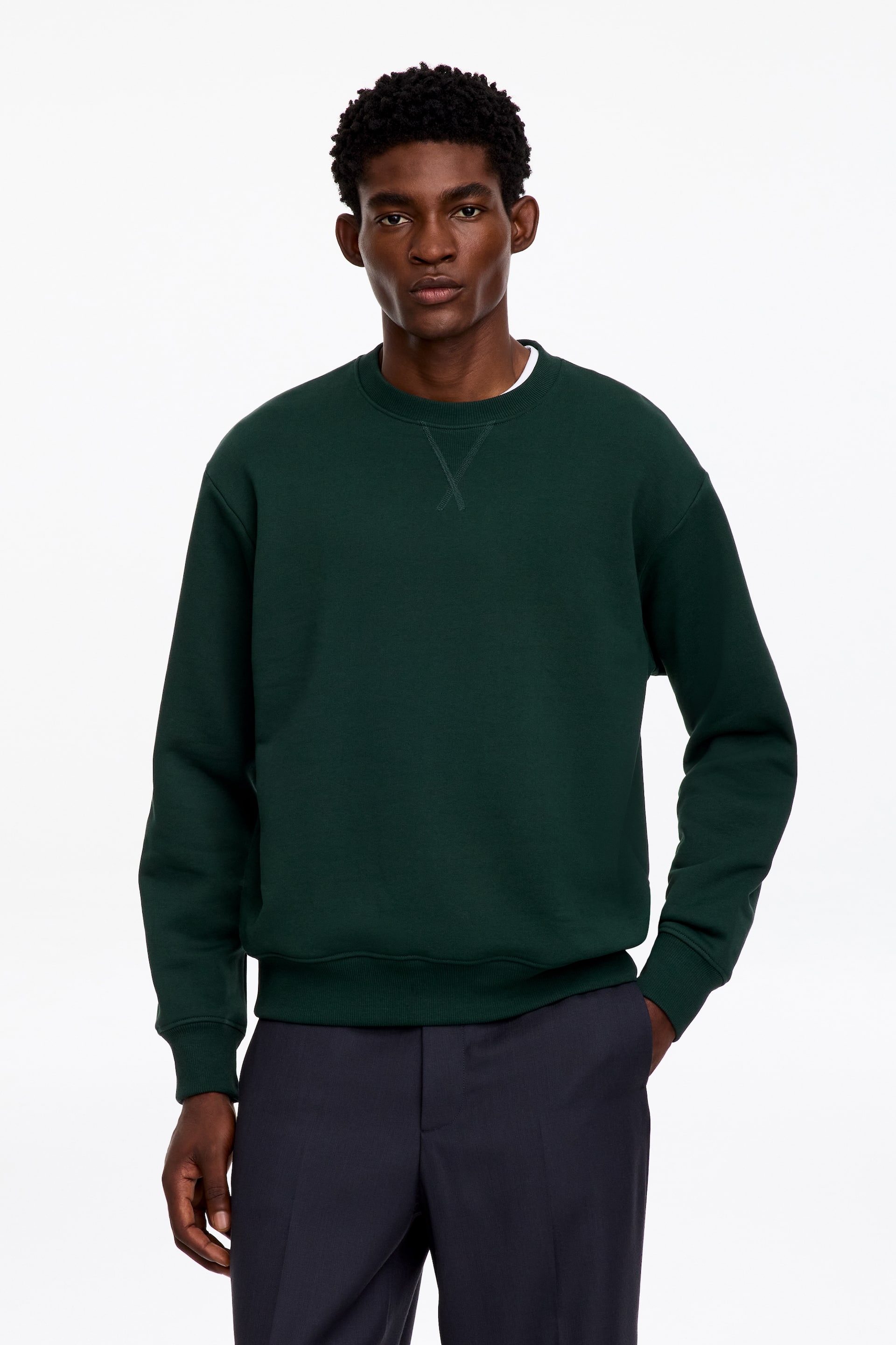 Agrandir l'image: Un sweat-shirt à col rond vert foncé est porté par le mannequin, avec un détail de surpiqûre en V visible à l’encolure, des poignets côtelés et un bord-côte à l’ourlet. Ce sweat-shirt structuré est porté sur un t-shirt blanc et associé à un pantalon foncé.