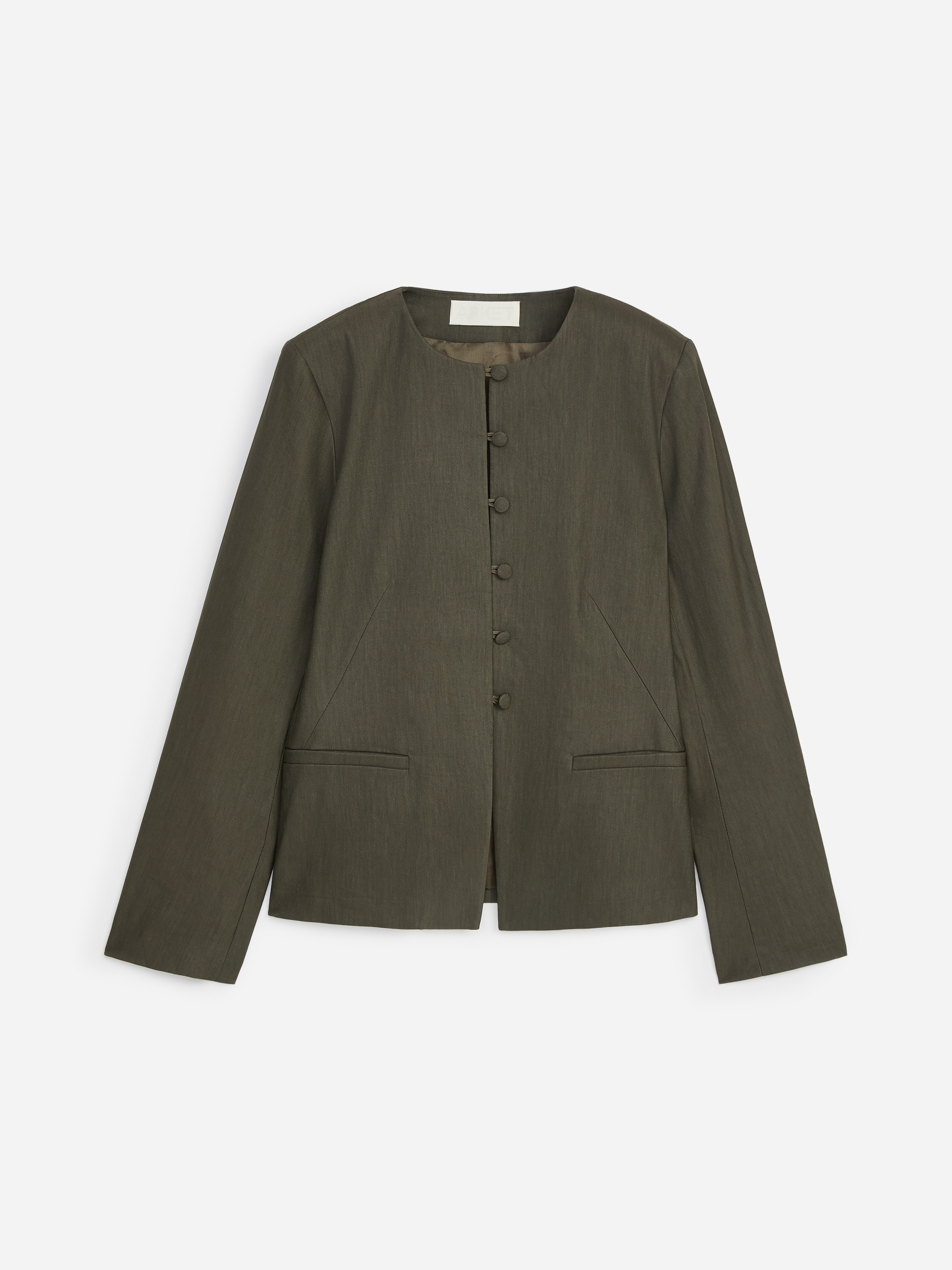 Modellen bÃ¤r en mÃ¶rkt olivgrÃ¶n kraglÃ¶s blazer, med knÃ¤ppning framtill med tygklÃ¤dda knappar och en ren silhuett. Blazern Ã¤r stylad Ã¶ver en vit topp och kombinerad med vida vita byxor.