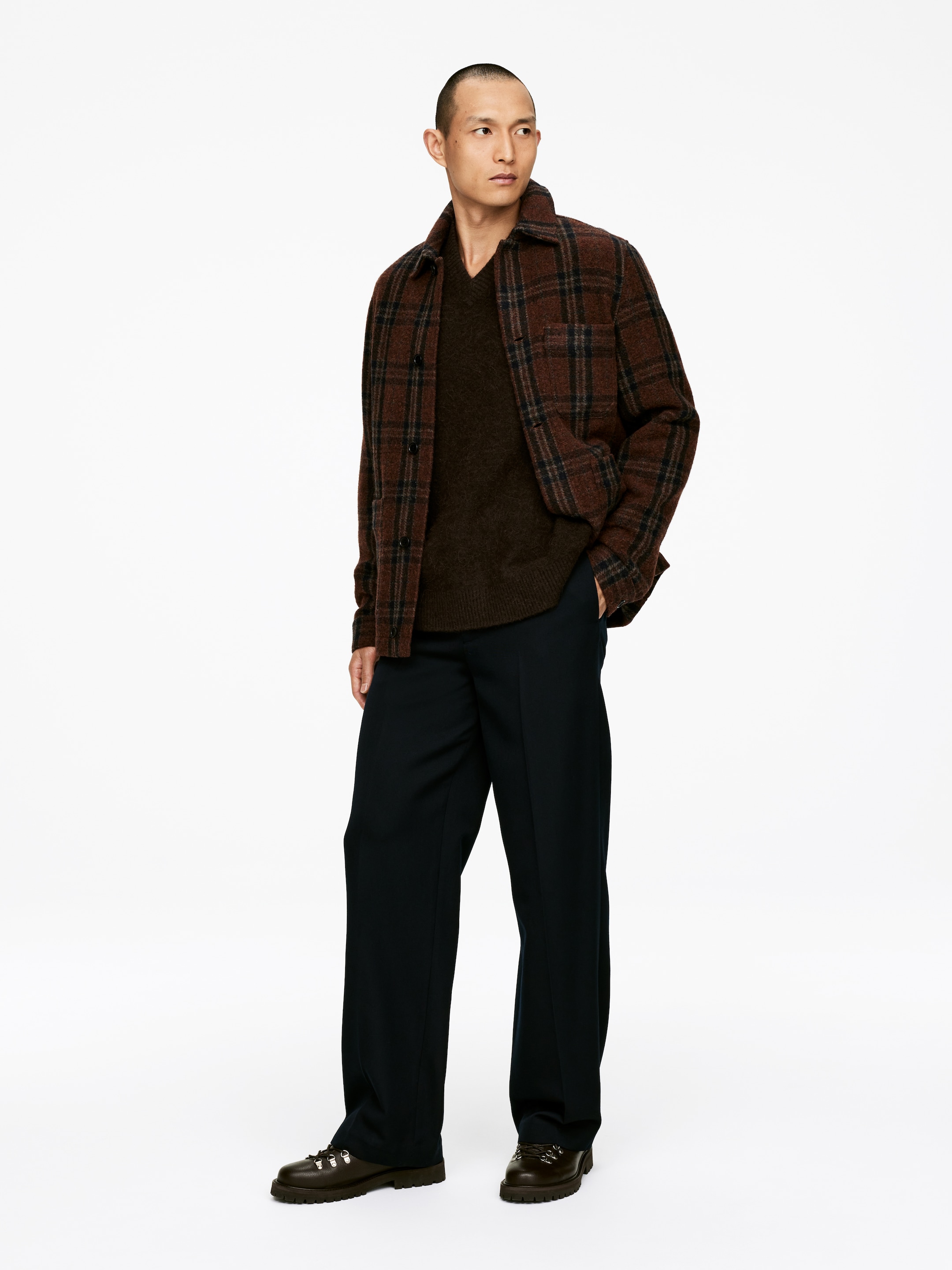 Agrandir l'image: Une veste-chemise à carreaux marron et noir, avec un col et une fermeture à boutons, est portée ouverte sur un pull à col en V marron foncé, offrant une coupe superposée décontractée. Un pantalon large bleu marine foncé est porté avec des bottes à lacets marron foncé qui ont des semelles épaisses.