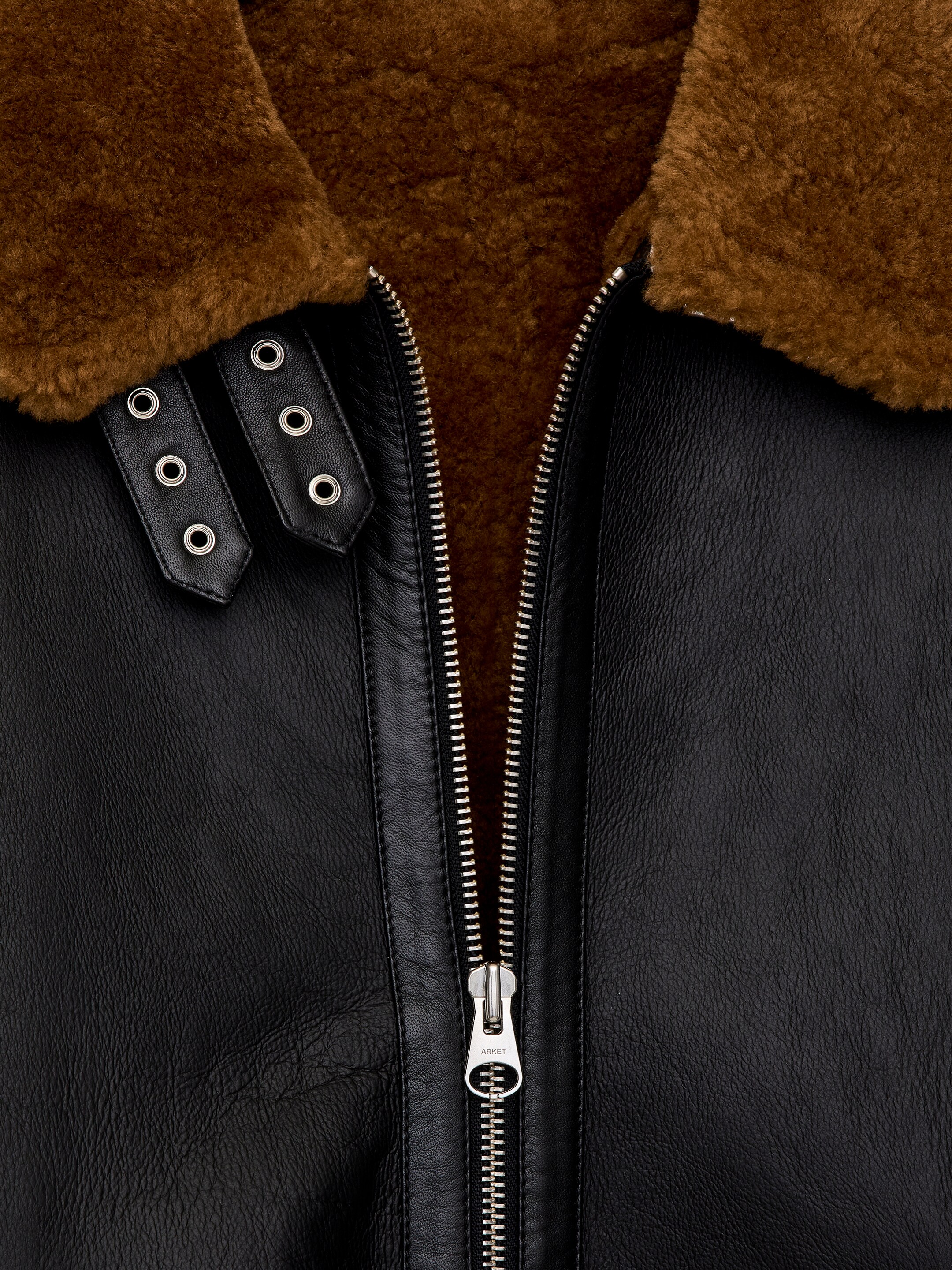 Vezi imaginea mai mare: PRIM-PLAN cu un sacou oversized din shearling, având un exterior din piele neagră și un guler de blană maro. Un fermoar argintiu cu deschidere în ambele sensuri străbate partea din față, parțial deschis și dezvăluind căptușeala de blană maro, în timp ce două curele reglabile din piele neagră cu catarame metalice argintii decorează gulerul.