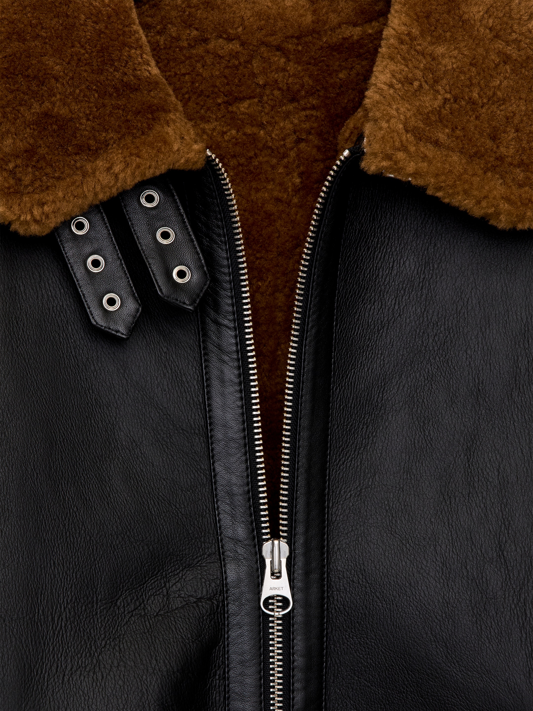 Agrandir l'image: GROS-PLAN d'une grande veste en shearling, avec un extérieur en cuir noir et un col en fourrure marron. Une fermeture éclair argentée à double sens descend sur le devant, partiellement ouverte et révélant la doublure en fourrure marron, tandis que deux sangles ajustables en cuir noir avec des boucles en métal argenté ornent le col.
