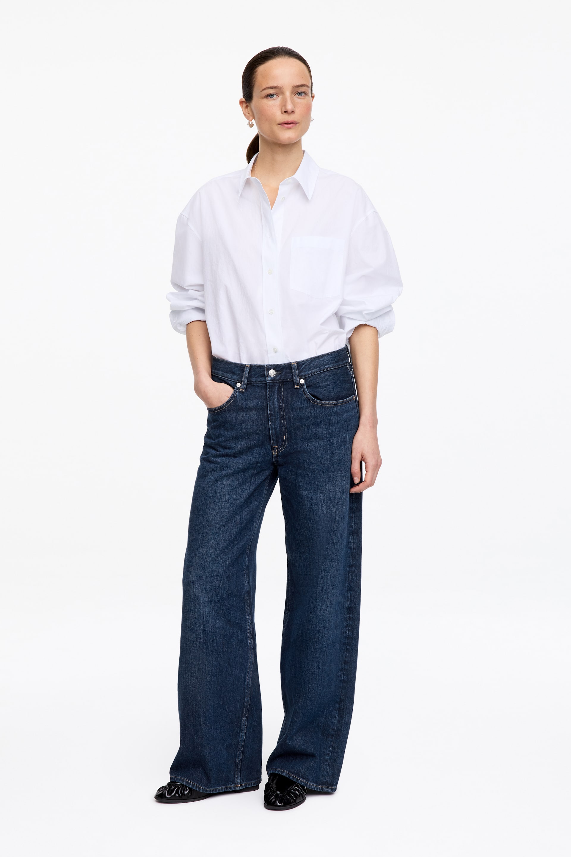 Agrandir l'image: Une chemise blanche à manches longues et boutonnée est présentée, face à l'avant, avec un col pointu, une patte de boutonnage à l'avant et une poche plaquée unique sur la poitrine gauche. Les manches semblent retroussées jusqu'aux avant-bras et la chemise est partiellement rentrée dans un jean bleu foncé, créant une coupe décontractée.