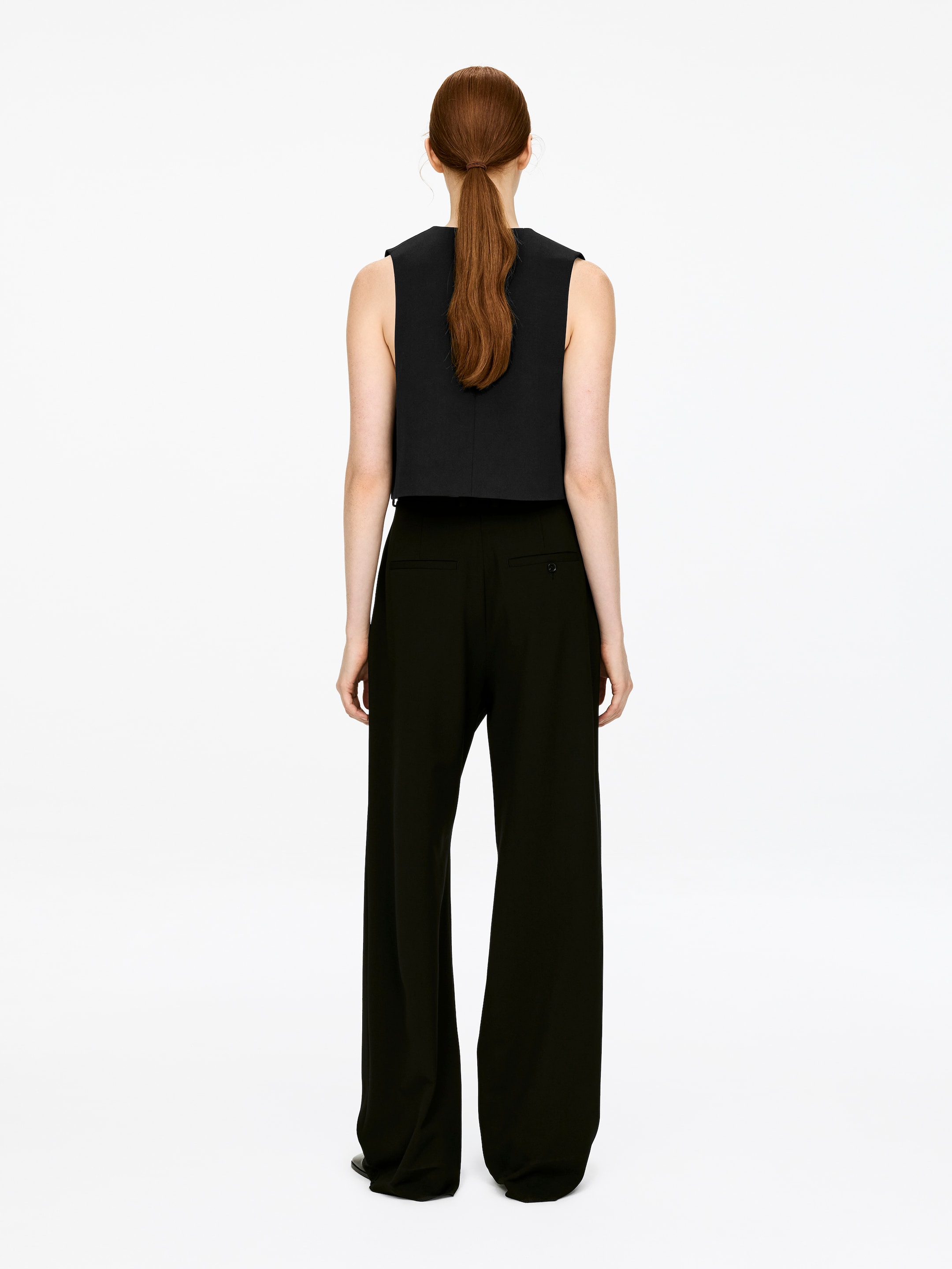 View larger image: A black, cropped, sleeveless tailored vest is being worn by the model, who is facing away from the viewer. The vest features a high back neckline and a smooth texture, and is being styled with dark, wide-leg trousers.