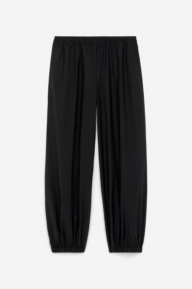 Schwarze Hosen werden aus einer Vorderansicht präsentiert, mit einer Barrel-Leg-Silhouette und einer entspannten Passform. Sie zeigen einen elastischen Bund, Seitentaschen und elastische Bündchen, die sich an den Knöcheln sammeln. - 1