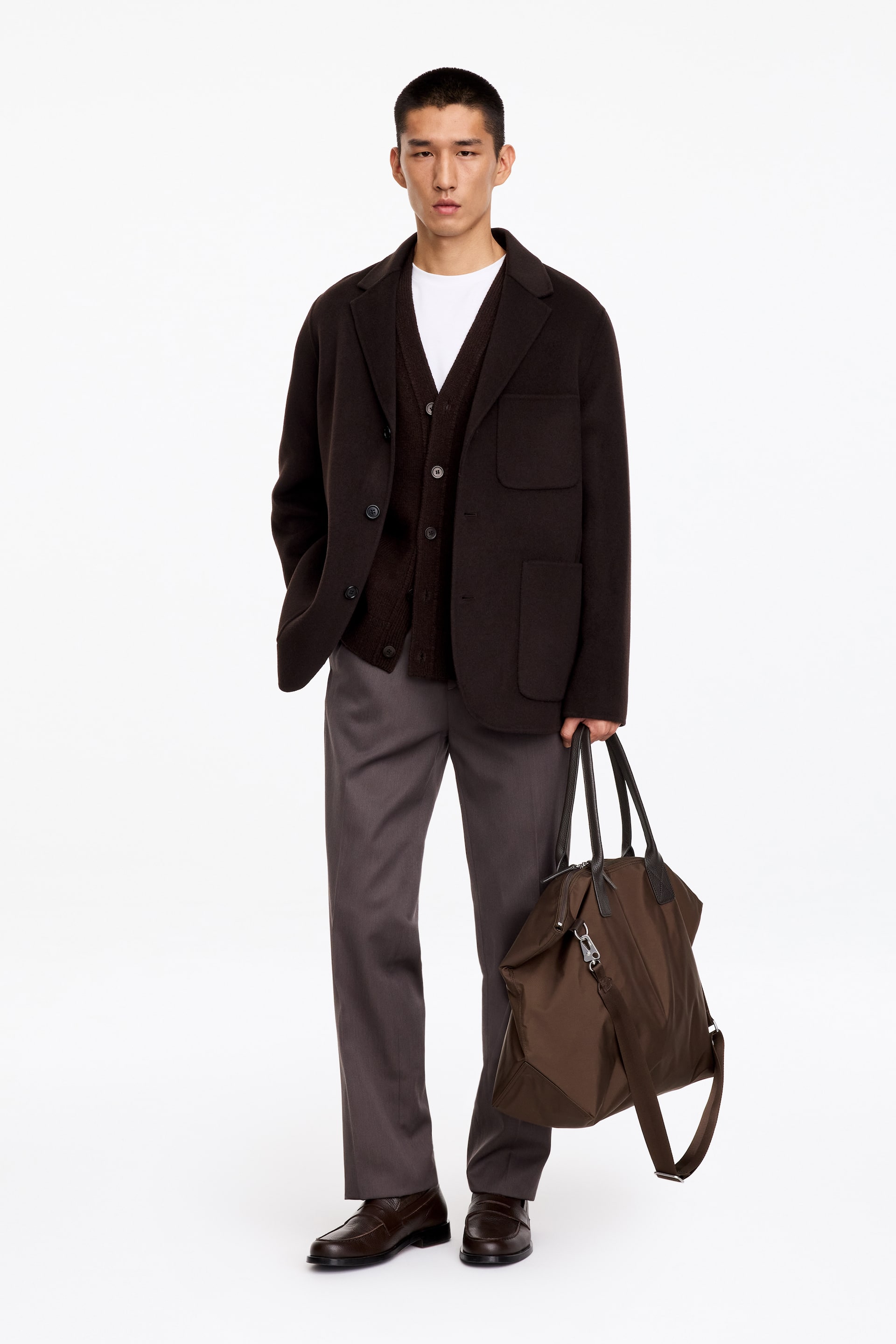Grösseres Bild ansehen: Eine braune Nylon-Tote-Bag mit schwarzen Ledergriffen und einem abnehmbaren Schulterriemen wird vom Model getragen, das sie an den oberen Griffen hält. Die Tasche verfügt über einen oberen Reissverschluss und silberfarbene Beschläge.