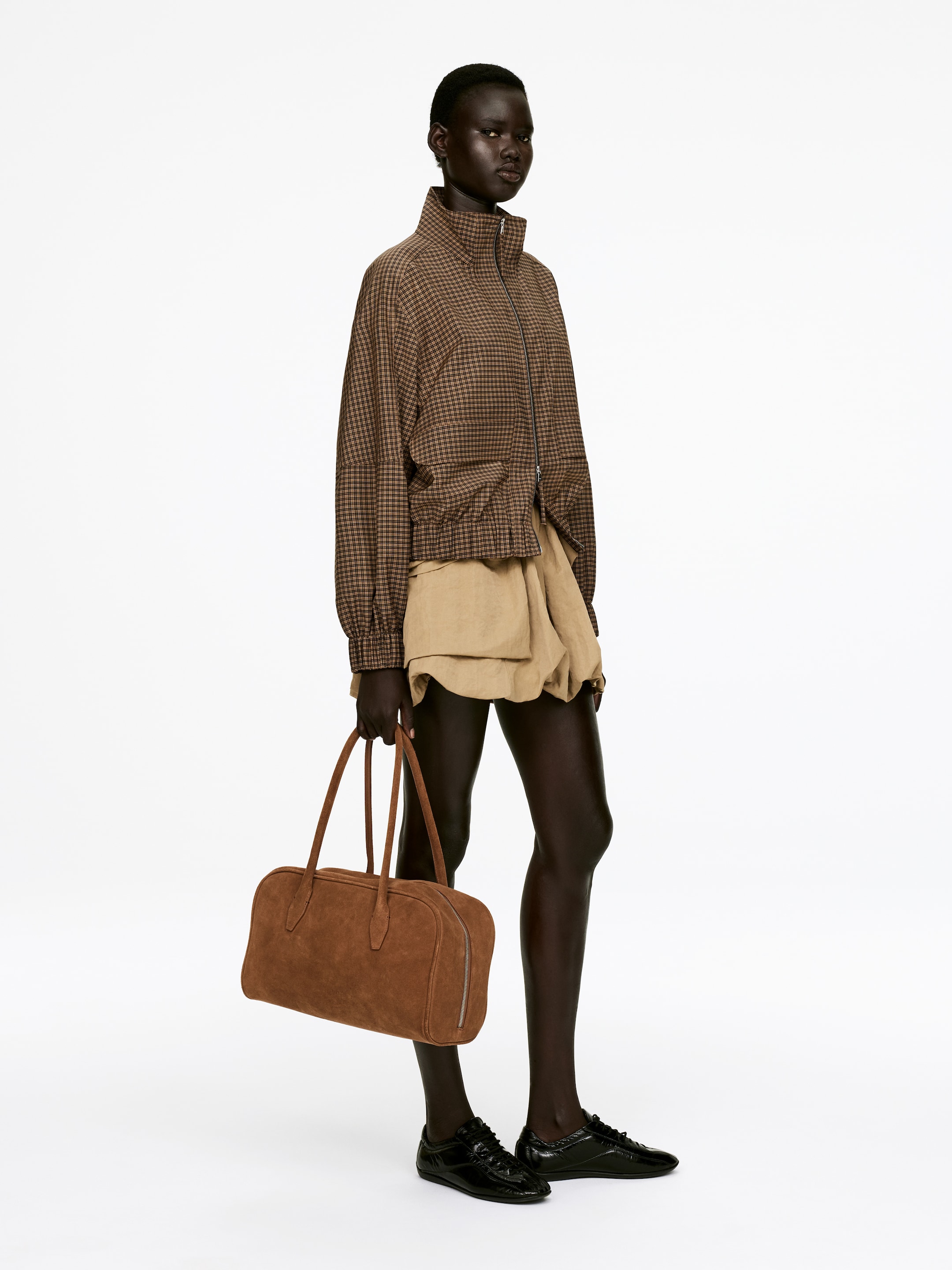 View larger image: A sporty jacket in a brown and black small check pattern is being worn by the model. It is featuring a stand-up collar, a two-way zip, and front welt pockets with flaps. The jacket's bottom and sleeve hems are elasticated, and it is layered over light brown shorts.