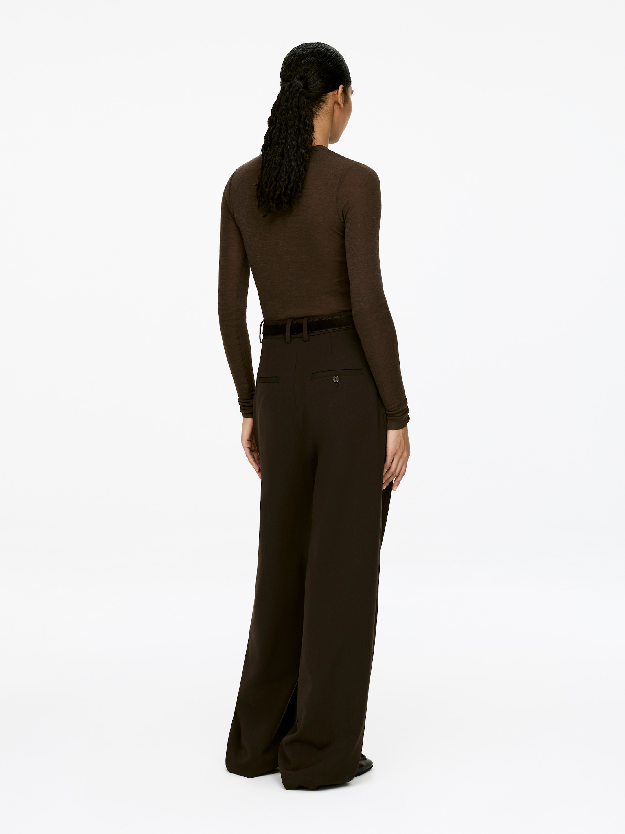 View larger image: A dark brown, tight-fitted merino wool top, featuring a slightly transparent appearance and long sleeves, is being worn by the model. The top is being tucked into dark brown trousers.