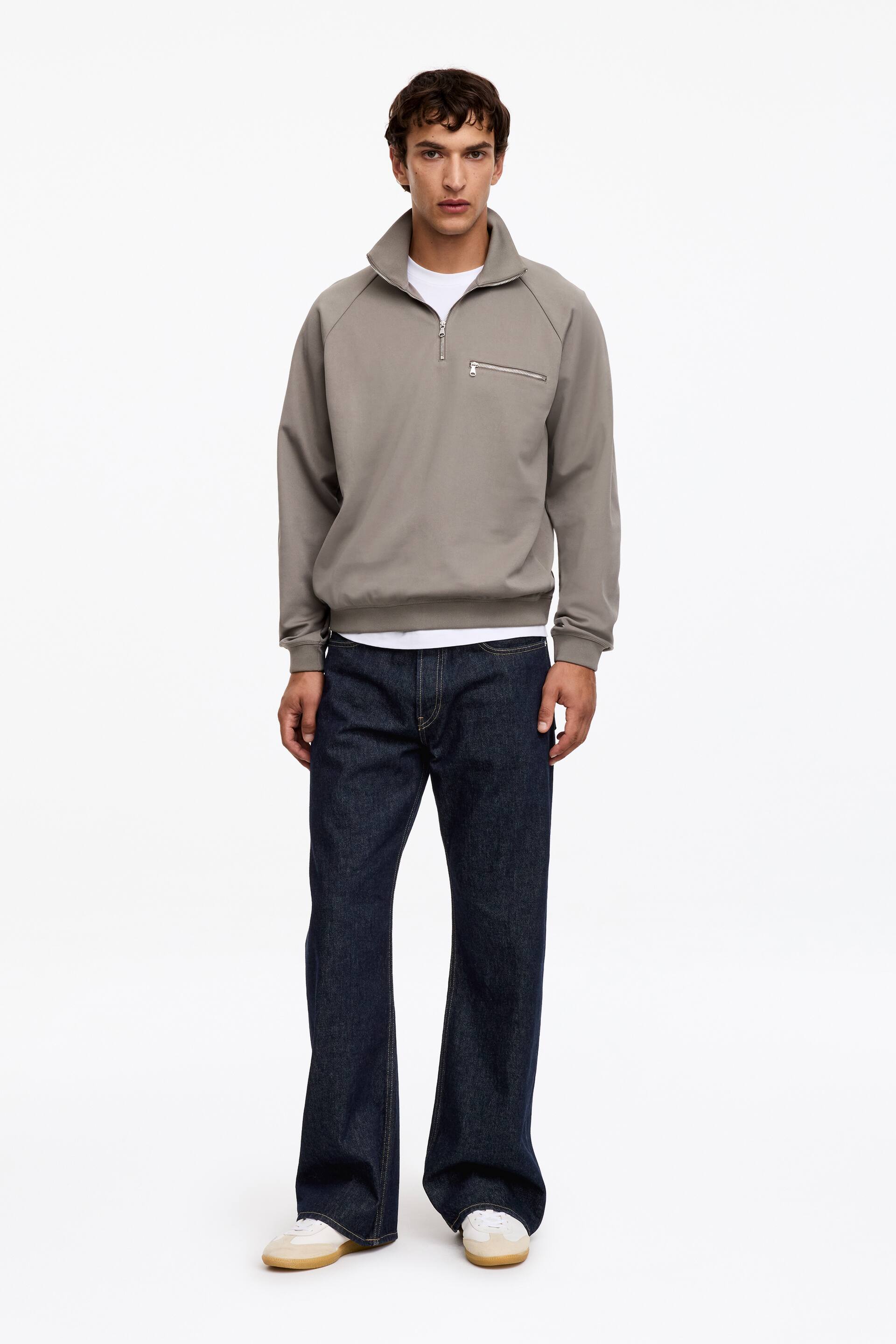 View larger image: A grey half-zip sweatshirt is being worn, facing forwards, featuring a high stand-up collar, raglan sleeves with ribbed cuffs, a ribbed hem, a silver-toned half-zip fastening, and a horizontal zip pocket on the left chest. It is layered over a white T-shirt and paired with dark blue wide-leg jeans.