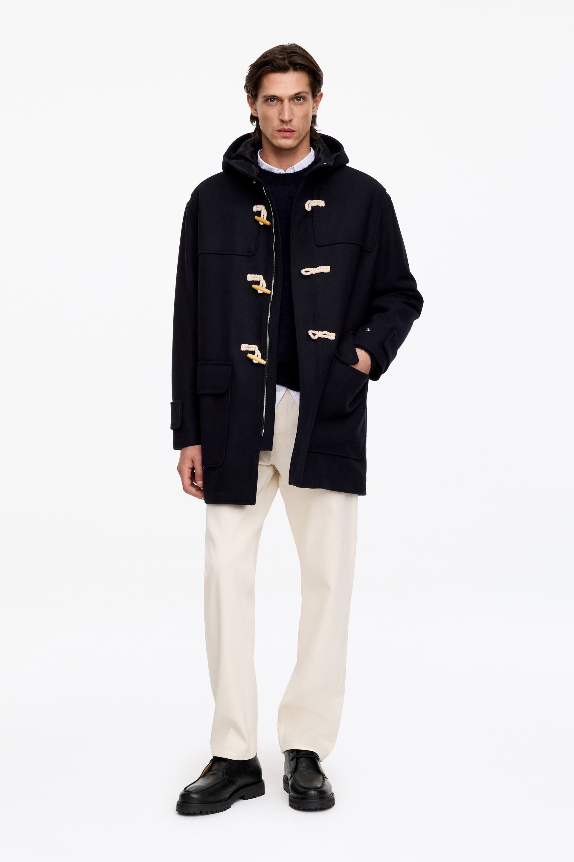 Agrandir l'image: Des bottines chukka montantes en cuir noir grainé, avec couture en plateau sur l’avant, lacets ton sur ton et épaisses semelles en caoutchouc noir, sont portées par le mannequin. Les bottines sont associées à un pantalon blanc cassé et à un duffle-coat à capuche bleu marine.