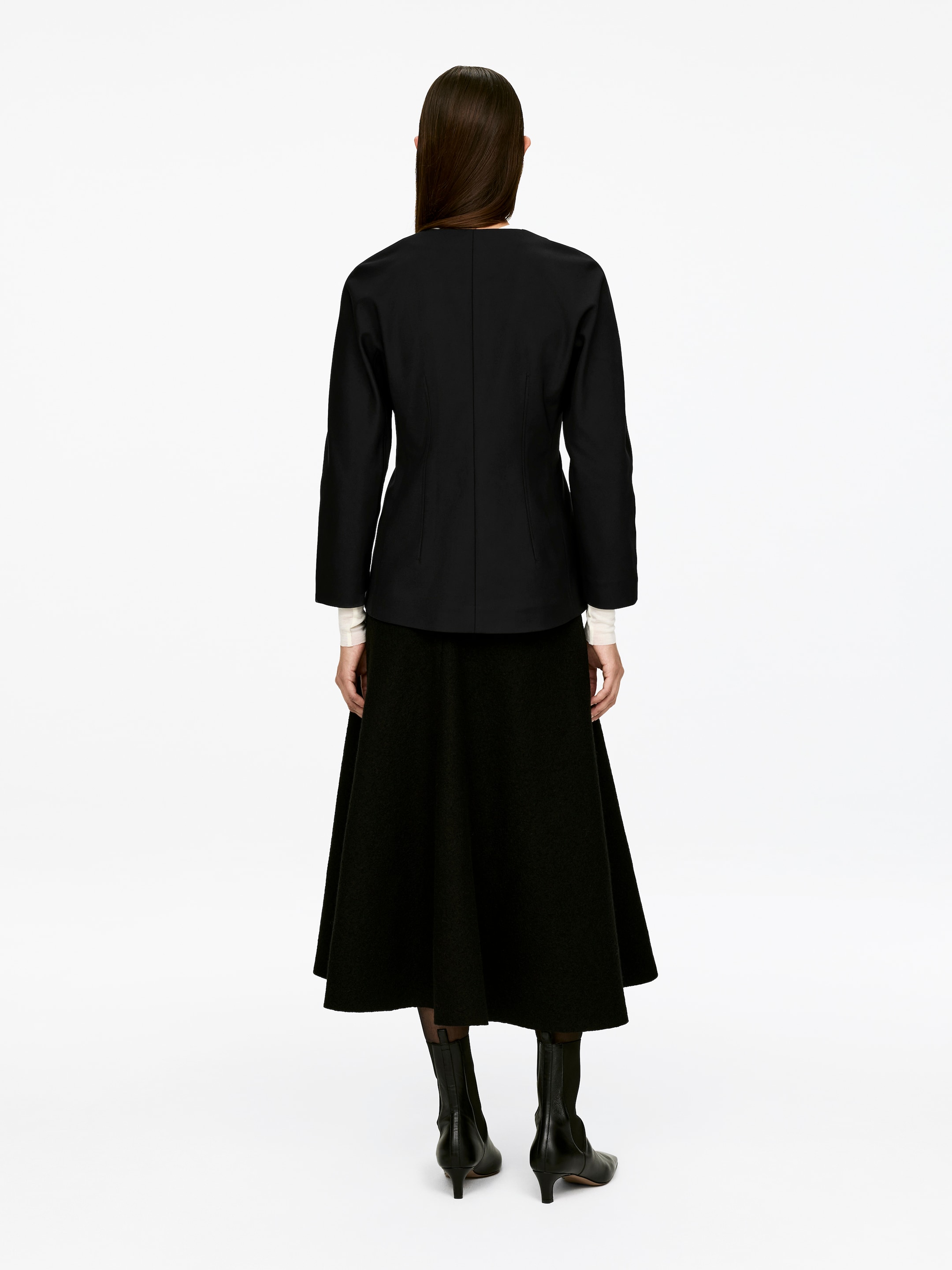 Visa större bild: En svart peplumtopp med en åtsittande midja och slank passform bärs av modellen, sedd bakifrån. Toppen har långa ärmar och en subtil peplumeffekt, och är lager på en vit långärmad topp och stylar med en svart midikjol och svarta stövlar.