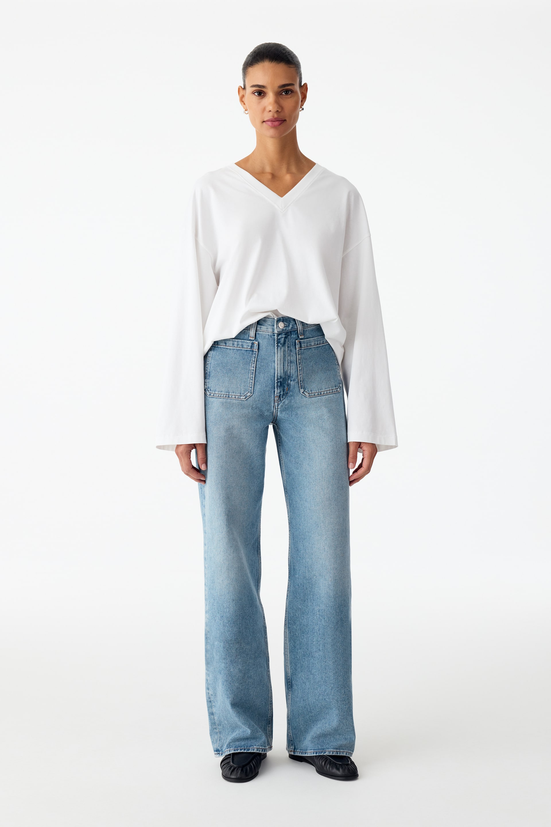 Visualizza immagine più grande: La modella indossa jeans in denim lavaggio azzurro chiaro con vita alta, tasche applicate frontali e gamba svasata. I jeans sono abbinati a un top bianco a maniche lunghe con scollo a V, parzialmente infilato nella vita.