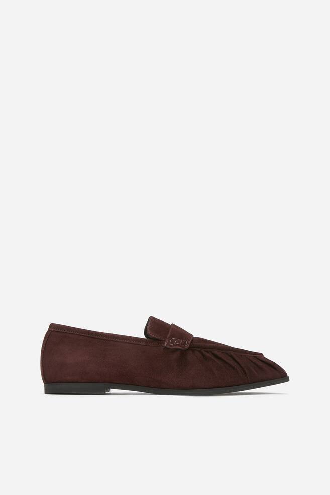 A dark brown suede loafer is facing left, featuring a pointed toe, a pleated vamp with a decorative strap across the top, and a thin, dark sole. - 1
