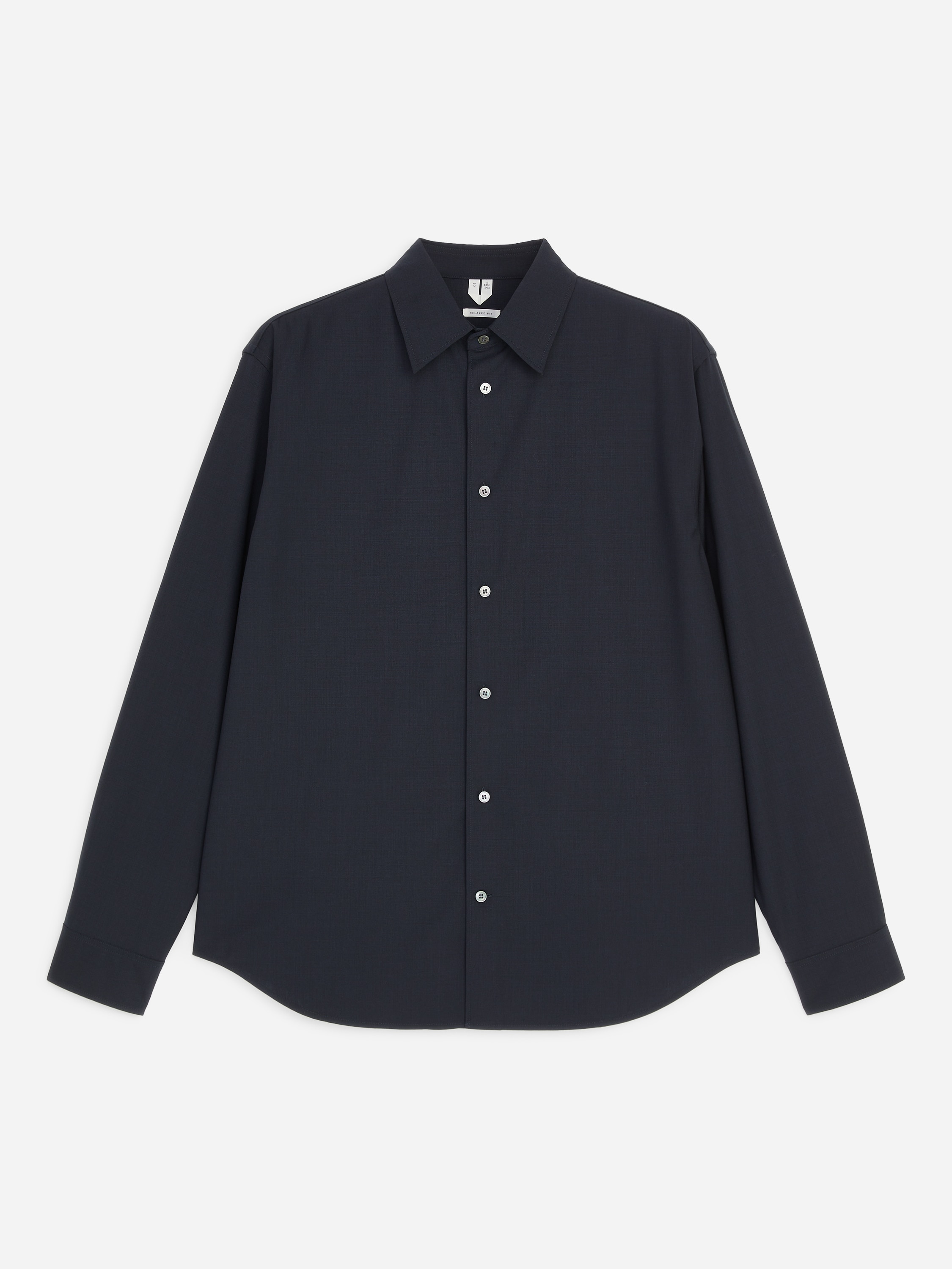 The model is wearing a dark grey, long-sleeved button-up shirt with a relaxed fit, a point collar and a curved hem. The shirt is styled untucked over dark trousers, with the modelâs hands resting in their pockets.