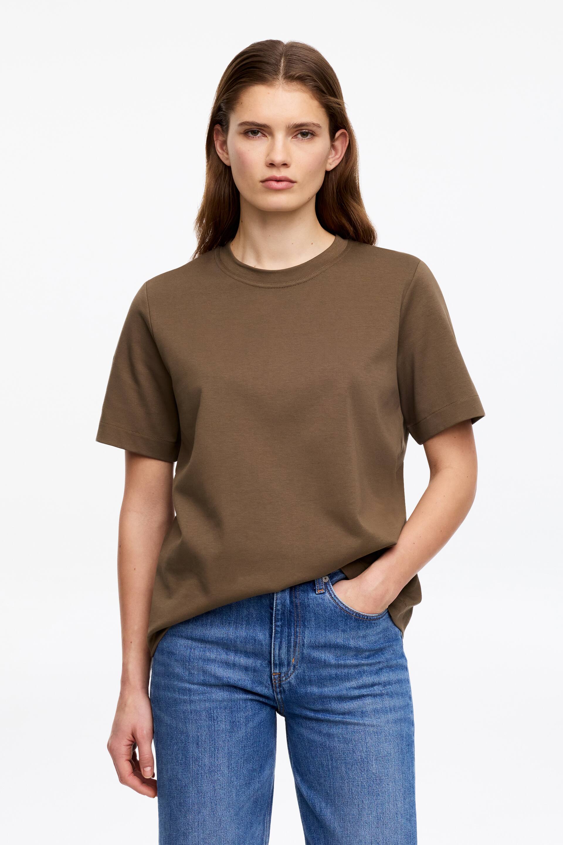 Visa större bild: En olivbrun t-shirt presenteras, framåtvänd, med rund hals, korta ärmar och en avslappnad passform. Dess fåll är nonchalant instoppad i ett par blå jeans på ena sidan, vilket framhäver dess fall.