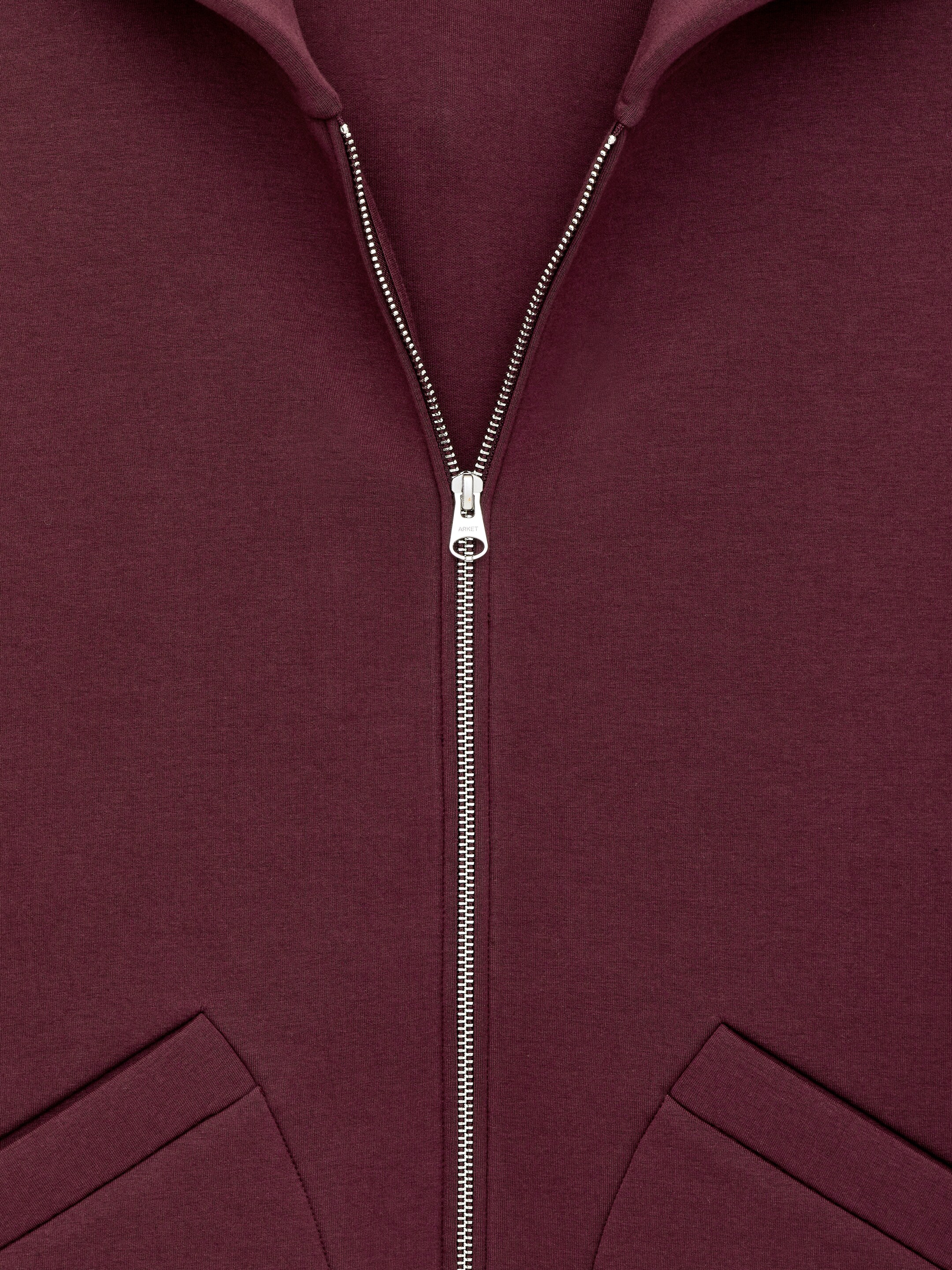 Grotere afbeelding bekijken: Een VOORAANZICHT van een donkerrode hoodie met rits, gemaakt van een gladde scuba-stof, heeft een zilverkleurige rits die in het midden naar beneden loopt, gedeeltelijk dichtgeritst. Twee schuine paspelzakken zijn zichtbaar aan de onderkant, en een capuchon van schelpstof omlijst de halslijn aan de bovenkant.