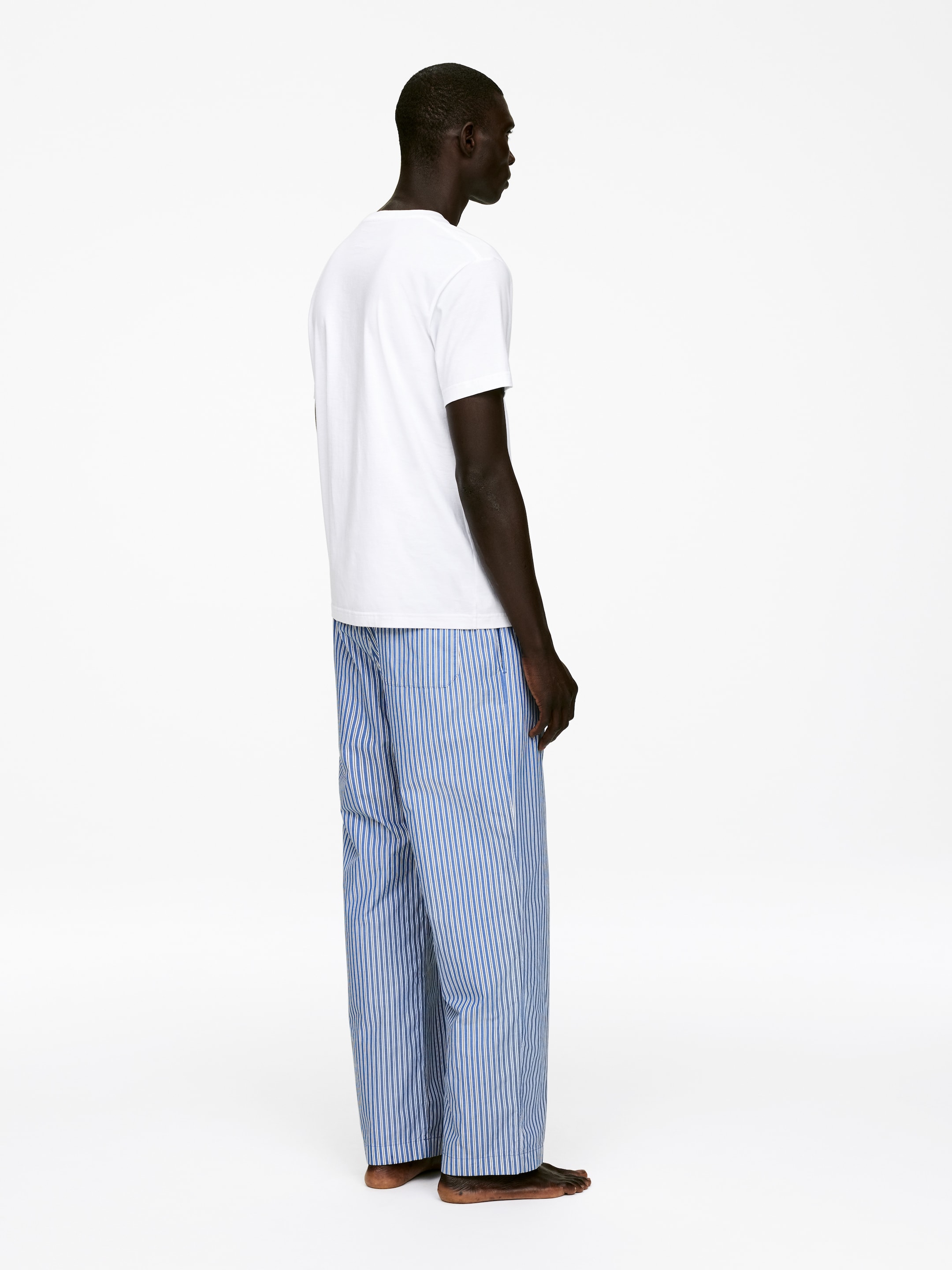 Agrandir l'image: Un t-shirt blanc à manches courtes est porté avec un pantalon à rayures verticales bleues et blanches, vu du côté droit. Le t-shirt est porté non rentré, et le pantalon présente une coupe ample et décontractée.
