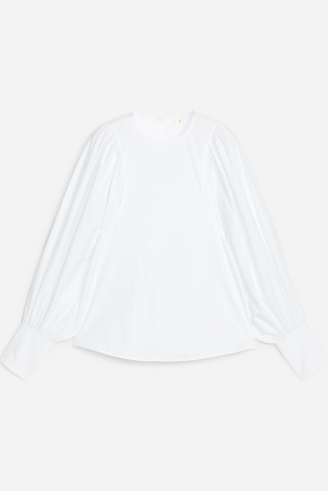 Een witte blouse wordt van voren getoond, met een ronde hals en lange gepofte mouwen die samenkomen in brede manchetten. De blouse eindigt met een subtiel gebogen zoom. - 1