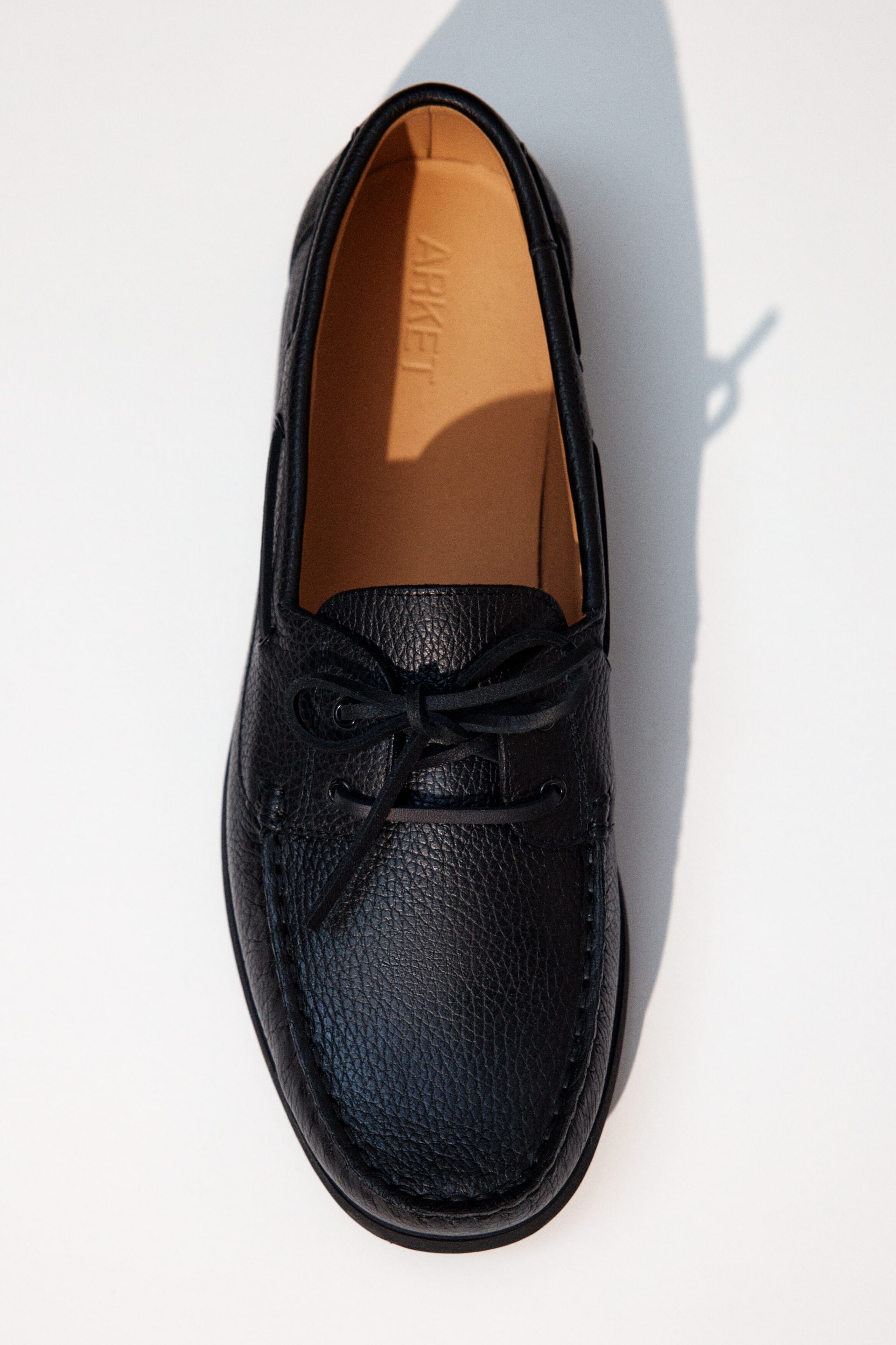 Größeres Bild anzeigen: Ein schwarzer Schuh aus genarbtem Leder liegt flach da und besticht durch eine Mokassin-ähnliche Zehennaht sowie schwarze Schnürsenkel, die zu einer Schleife gebunden sind. Die hellbraune Innensohle des Schuhs ist sichtbar und zeigt den Markennamen 'ARKET'.