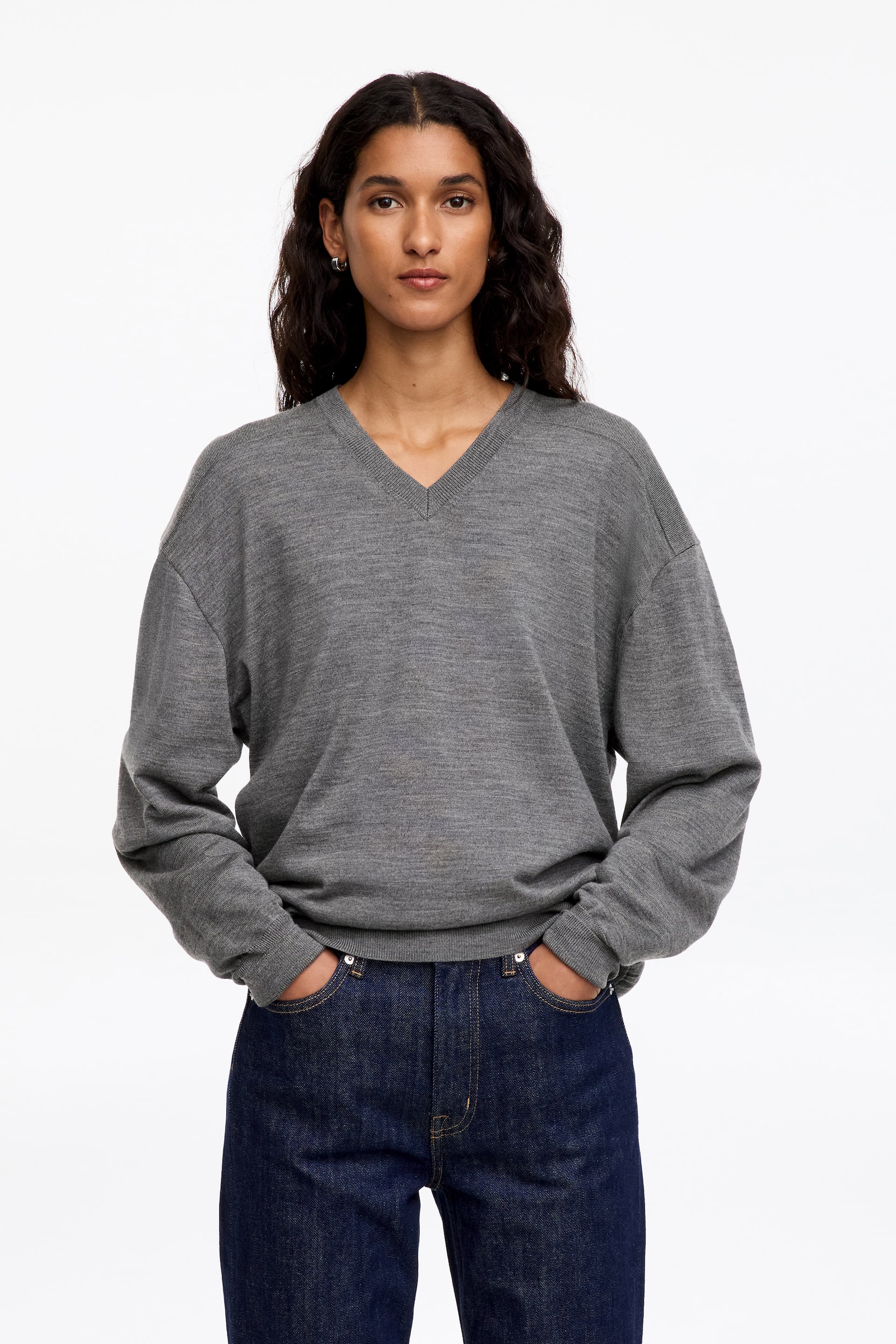 Agrandir l'image: Un pull gris en maille fine à col en V est porté par le mannequin, qui se tient debout, les mains dans les poches d’un jean bleu foncé. Le pull présente une coupe décontractée avec des épaules tombantes et des finitions côtelées au niveau de l’encolure, des poignets et de l’ourlet.