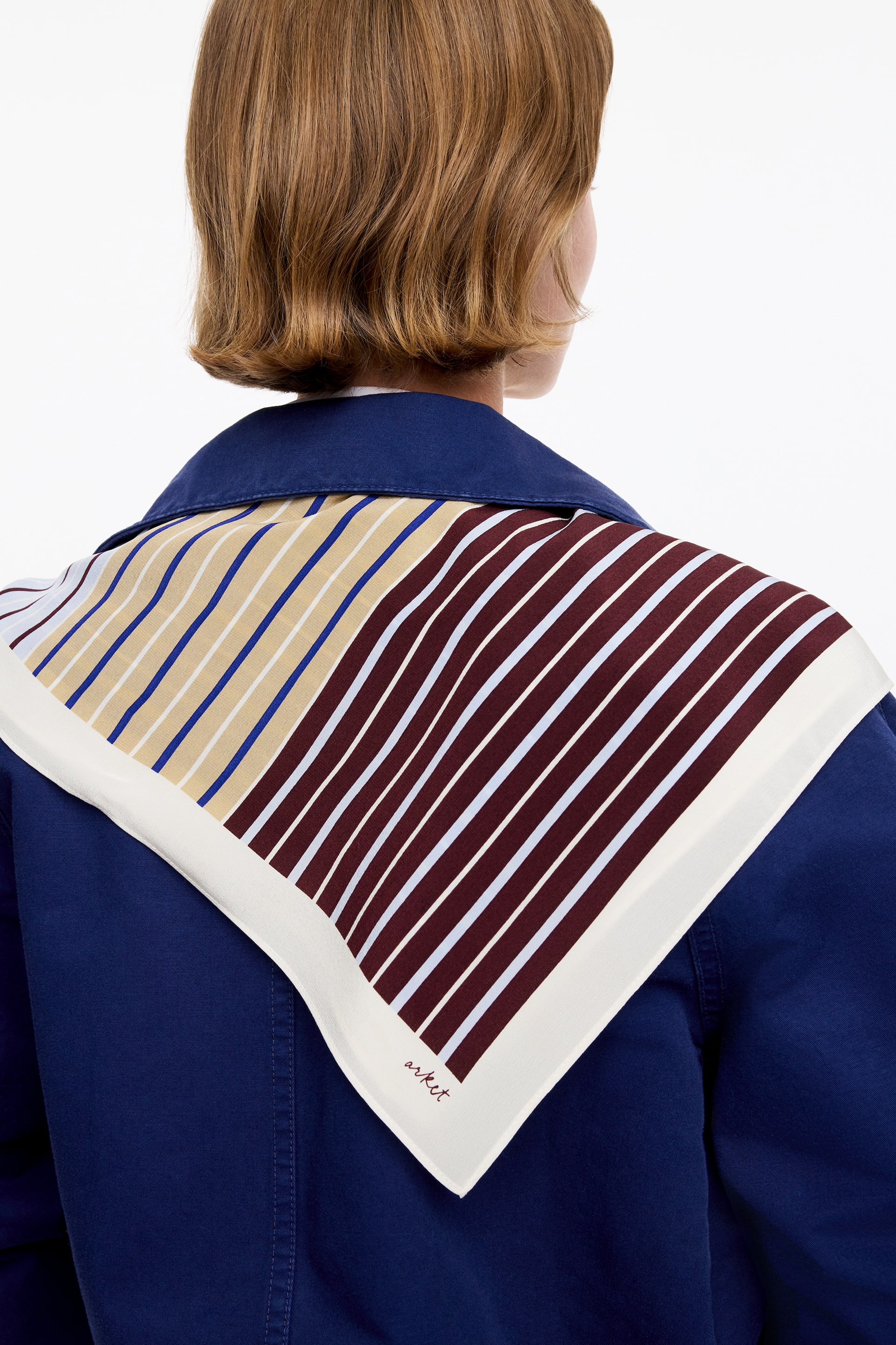 View larger image: The square scarf is draped over the shoulders of a navy jacket, visible from the back. It features diagonal stripes in maroon and white on one side, and tan, blue, and white on the other, all framed by a cream border with "arket" visible.