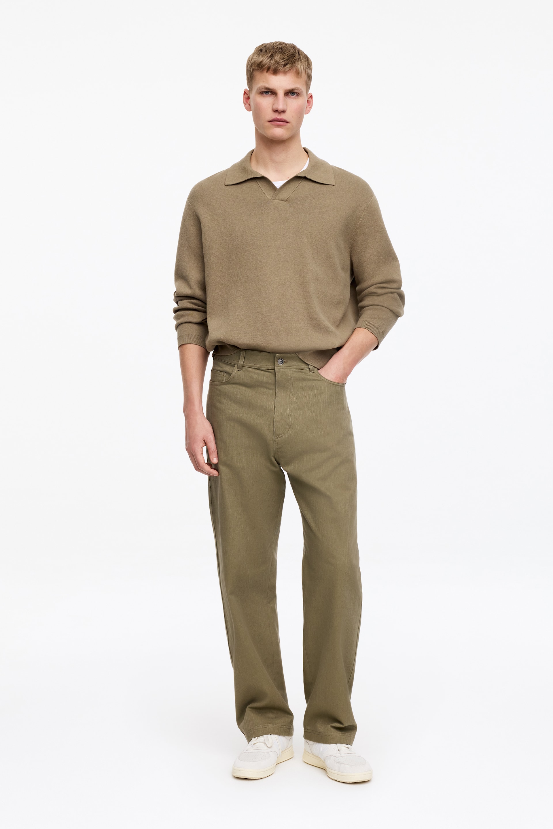 Zobrazit větší obrázek: Model má na sobě klasické olivově zelené kalhoty z bavlněného kepru s volnějším střihem, nohavicemi s přímým střihem, pětikapsovým provedením a viditelným knoflíkovým zapínáním nad zipem. Jeho ruka spočívá v přední kapse kalhot.