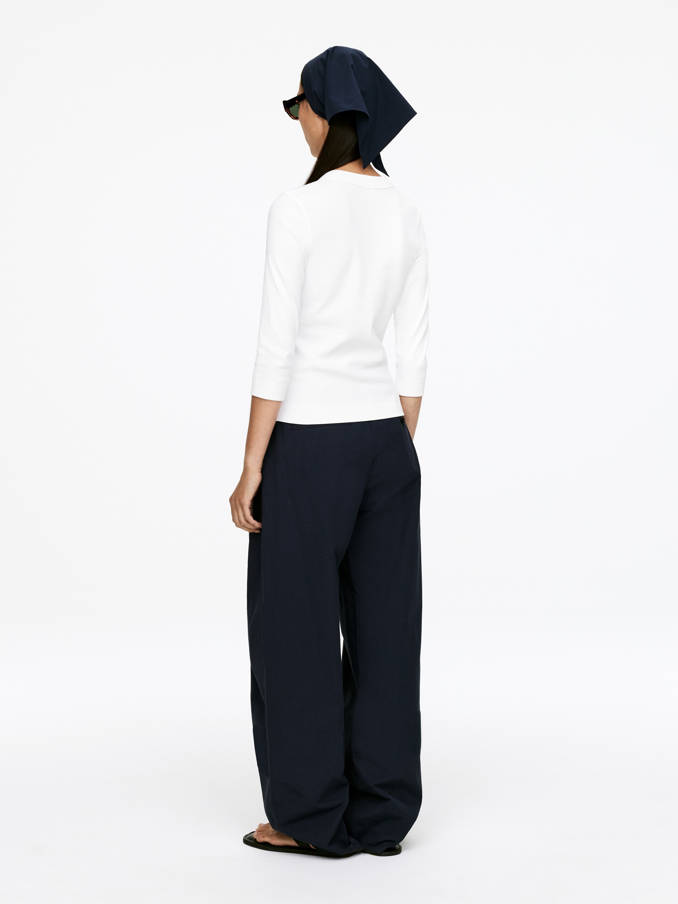 Agrandir l'image: Un haut blanc ajusté à manches trois-quarts, avec un décolleté arrondi dans le dos, est porté avec un pantalon sombre, ample et à jambes larges qui tombe jusqu'au sol, le tout vu de dos.