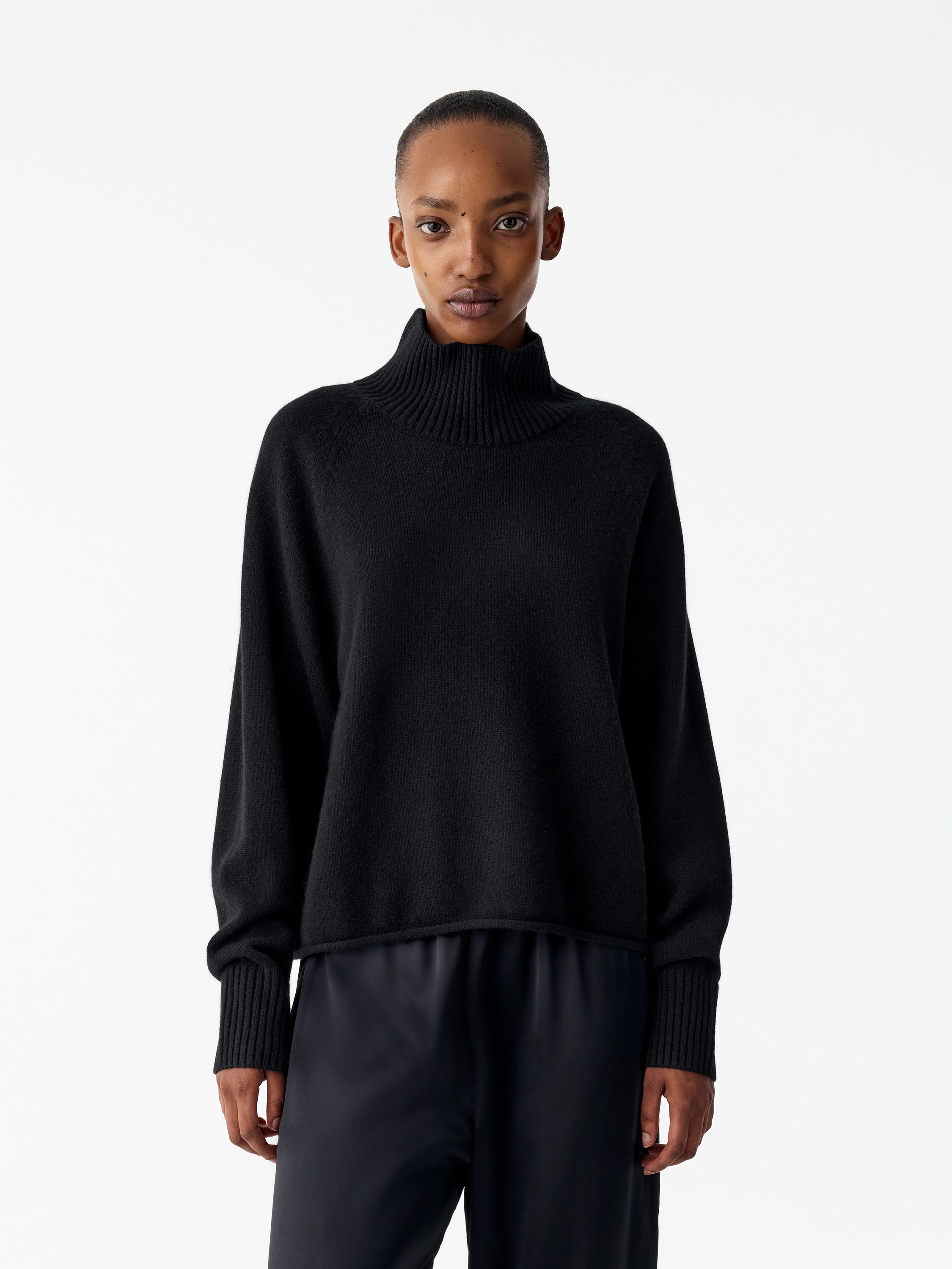 Un maglione nero in maglia è mostrato frontalmente, con un collo alto finto a coste e maniche lunghe con ampi polsini a coste. Il maglione presenta una silhouette rilassata e squadrata, con un orlo pulito leggermente arrotolato.
