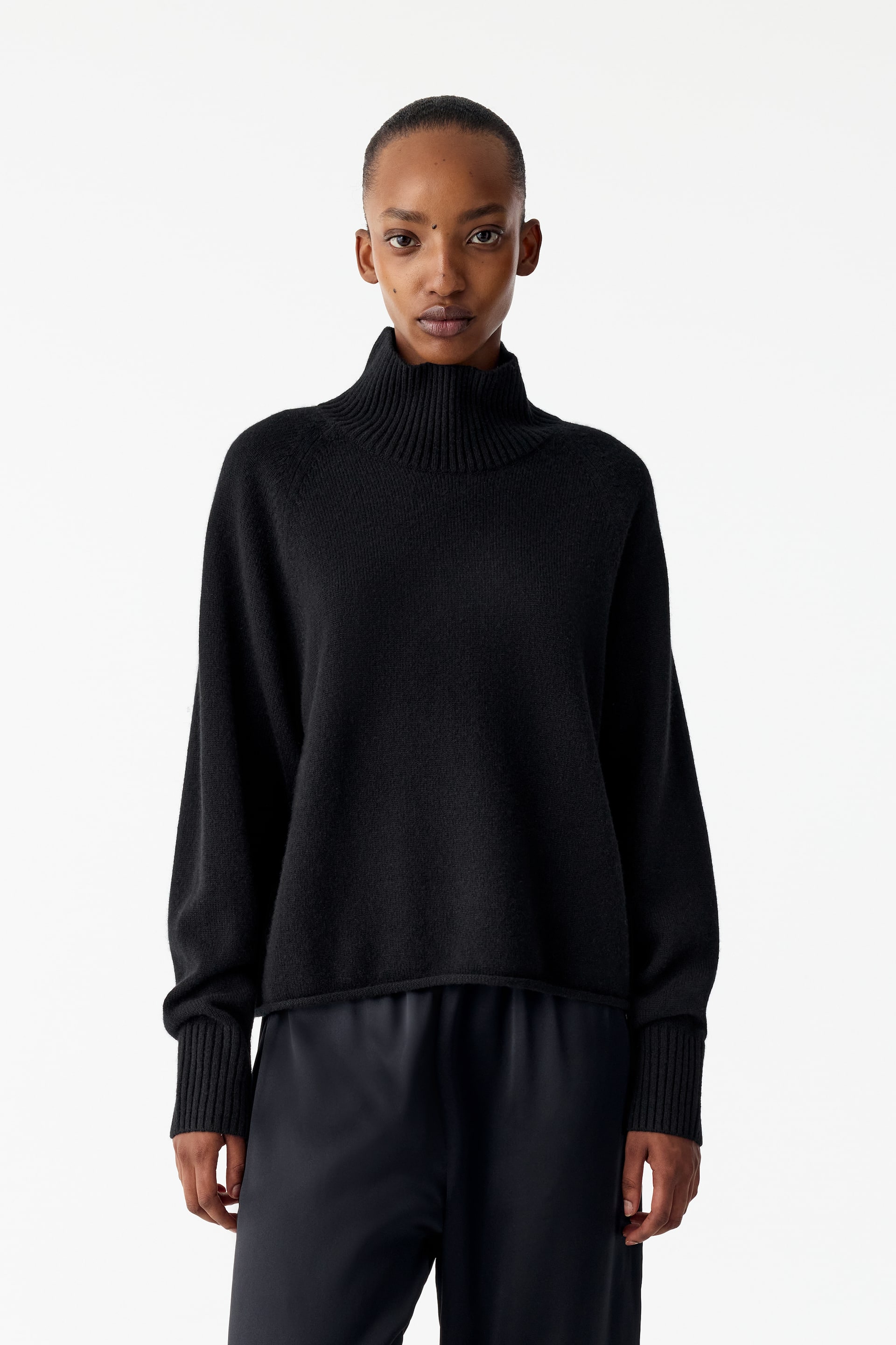 View larger image: A black cashmere roll-neck jumper featuring a high ribbed collar, raglan sleeves, and long ribbed cuffs is being worn by the model. The jumper has a relaxed silhouette and is paired with dark, loose-fitting trousers.