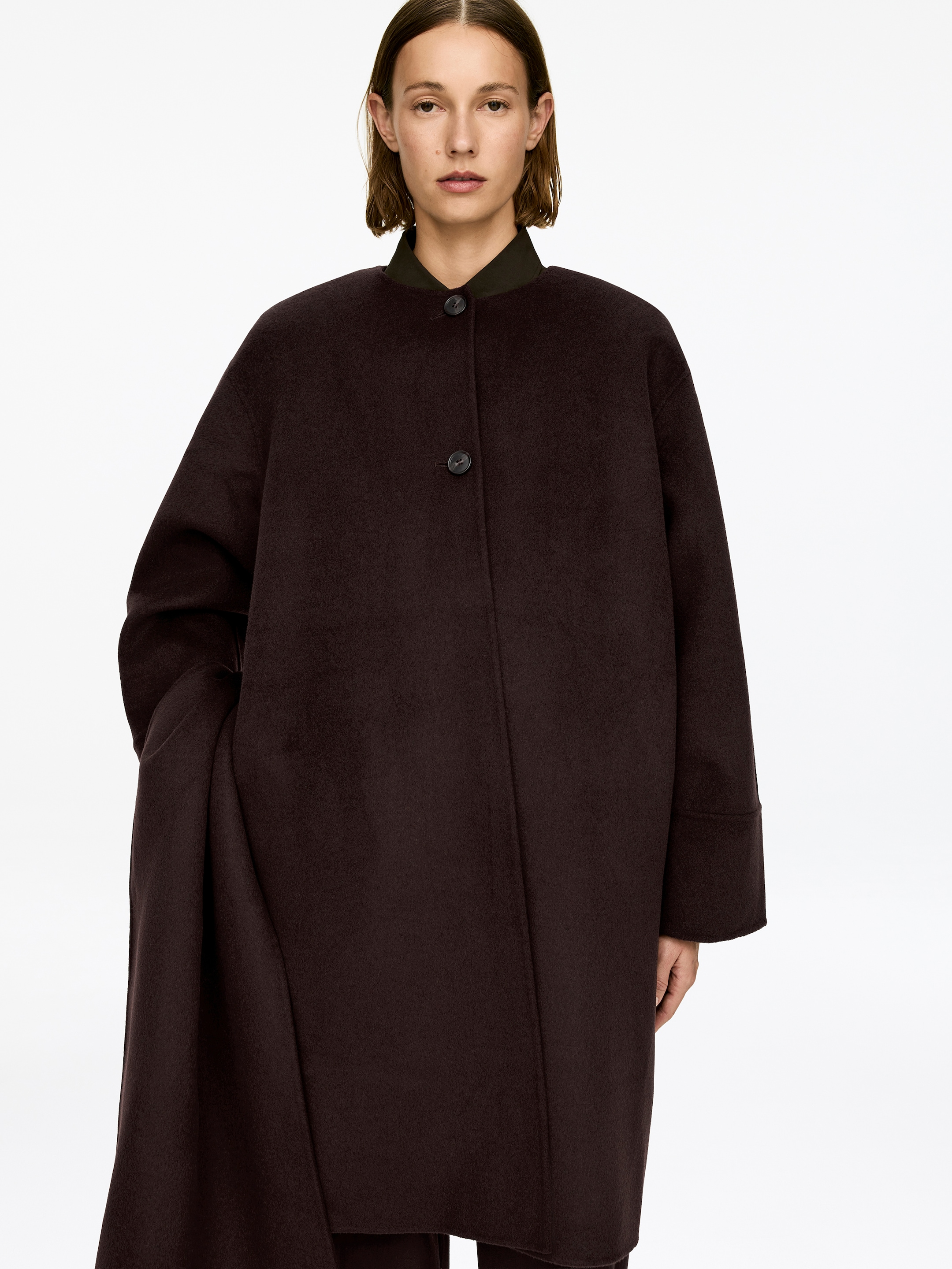 Agrandir l'image: Un manteau sombre en mélange de laine à double face avec un col bande est porté par le mannequin, comportant une fermeture à boutons avec deux boutons visibles. Il comprend des poignets hauts, de profondes fentes latérales et un détail de cape d'un côté. Le mannequin place une main dans une poche latérale, et une chemise sombre à col est visible sous le manteau.