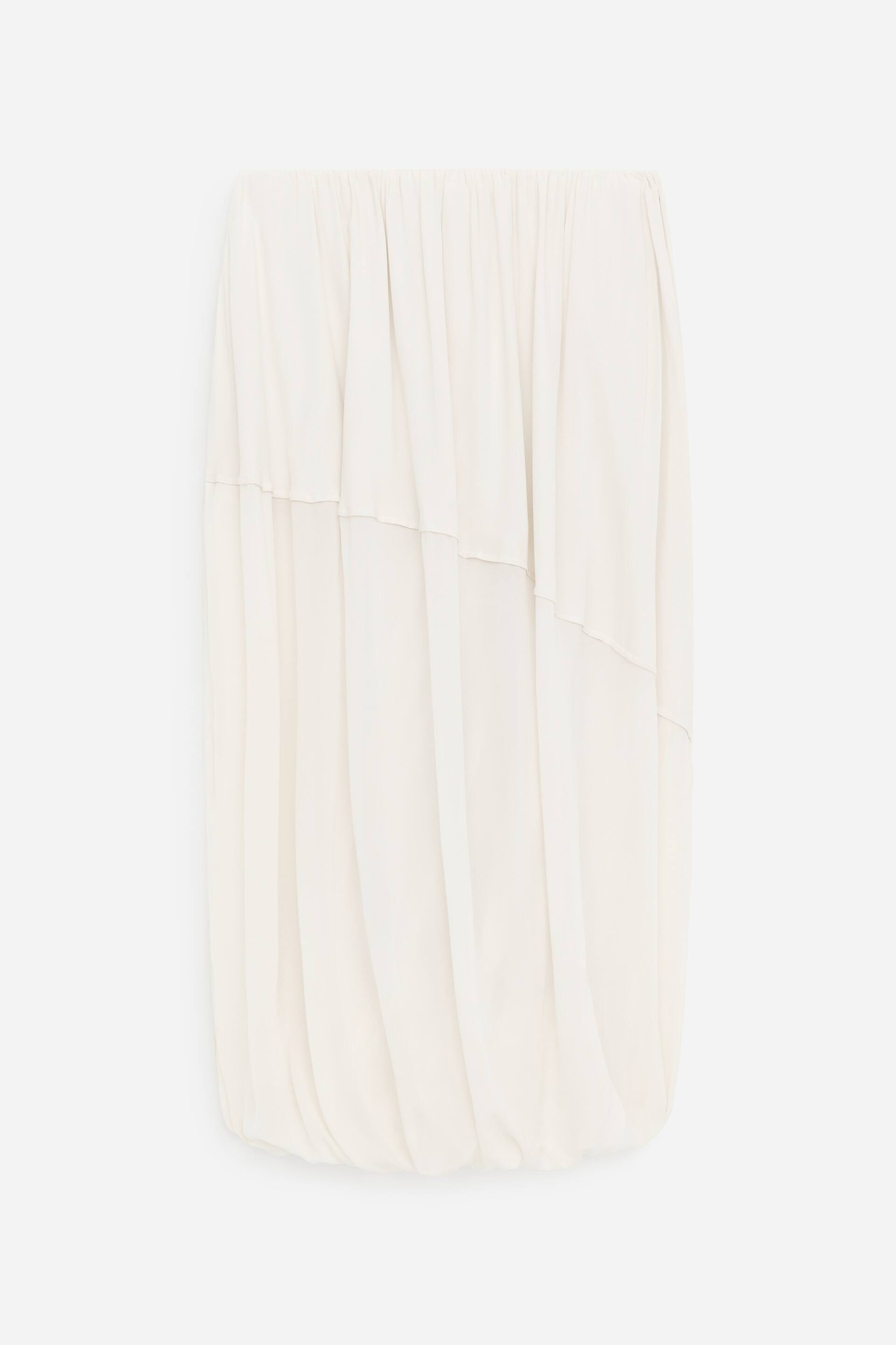 Visa större bild: En off-white kjol visas från framsidan, med en voluminös, bubbelstil silhuett. Den har en rynkad elastisk midja, flödande tyg som bildar vertikala draperingar, och horisontella sömlinjer som skapar en lager-på-lager-effekt över framsidan.