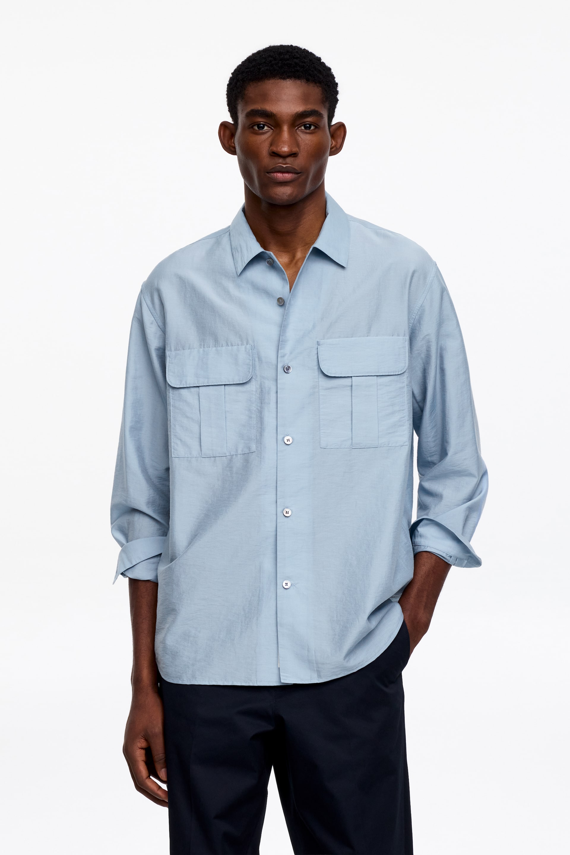 Se større billede: Den lyseblå skjorte med knapper har en spids krave, en fuld knaplukning med lyse knapper og to store brystlommer med flap, hver med lodrette læg. Dens lange ærmer er rullet op til underarmene, og den har en afslappet pasform med lige kant, båret uden at være stoppet ind.