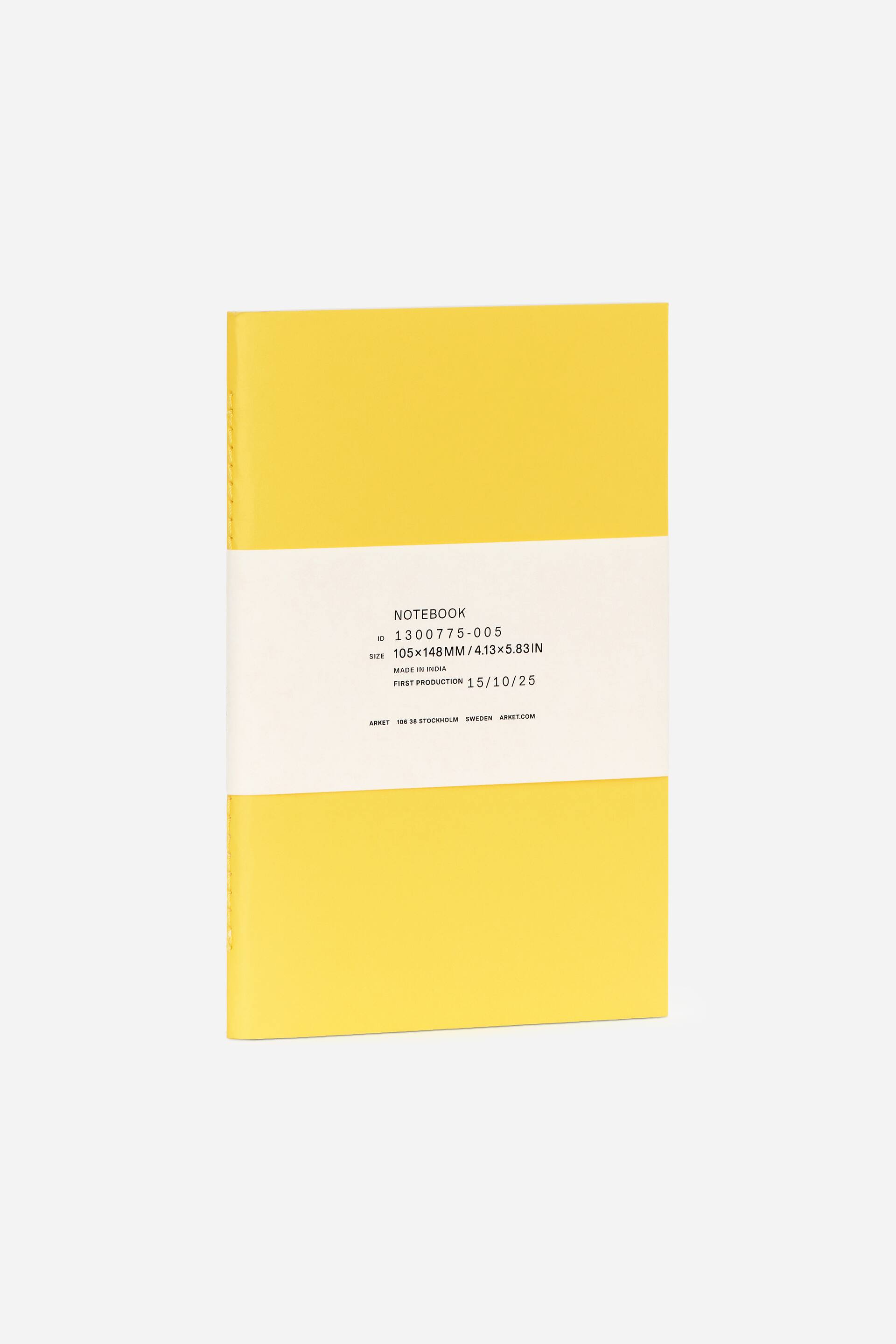 Se større billede: En gul notesbog præsenterer sin forside, let vinklet for at afsløre sin syede ryg på venstre side. Et hvidt papirbånd omkranser horisontalt midten og viser produktdetaljer, herunder dens 105x148MM (A6) størrelse i sort tekst.