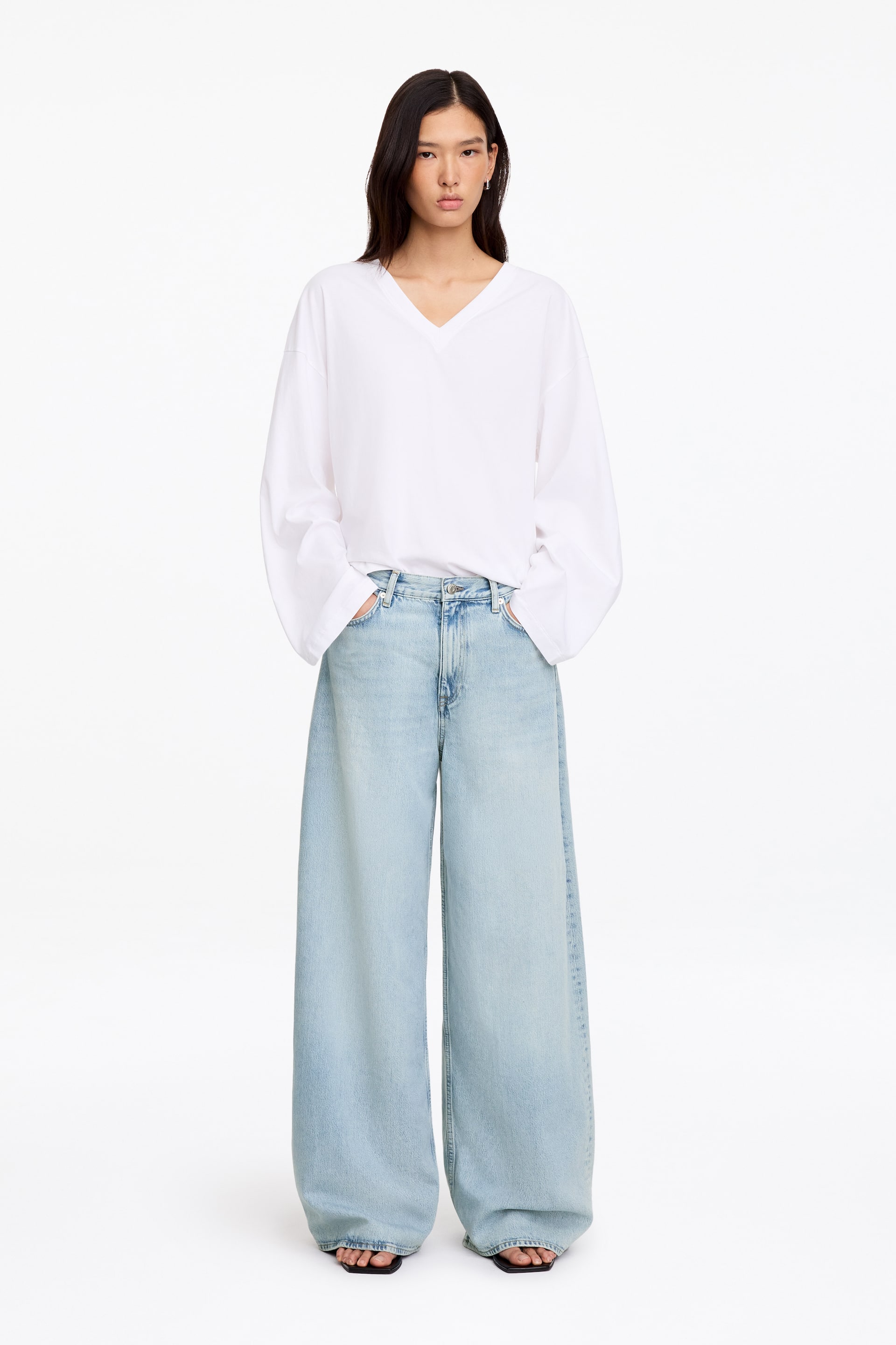 Visualizza immagine più grande: La modella indossa un top bianco con scollo a V e maniche lunghe e jeans in denim lavaggio chiaro a gamba larga. La modella è in piedi con le mani inserite nelle tasche anteriori dei jeans, mettendone in evidenza la vestibilità rilassata e ampia.