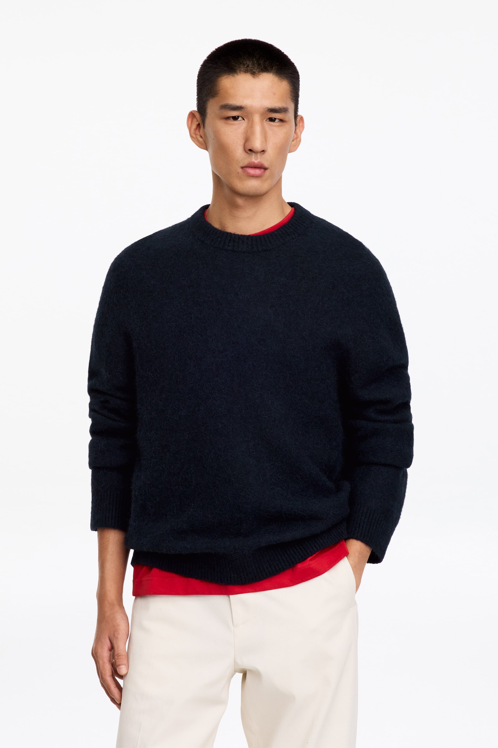 Visa större bild: En marinblå jumper med rund hals i avslappad passform bärs av modellen ovanpå en röd T-shirt och benvita byxor. Jumpern har en texturerad, mjukstickad konstruktion med ribbade detaljer vid krage, ärmslut och nederkant.