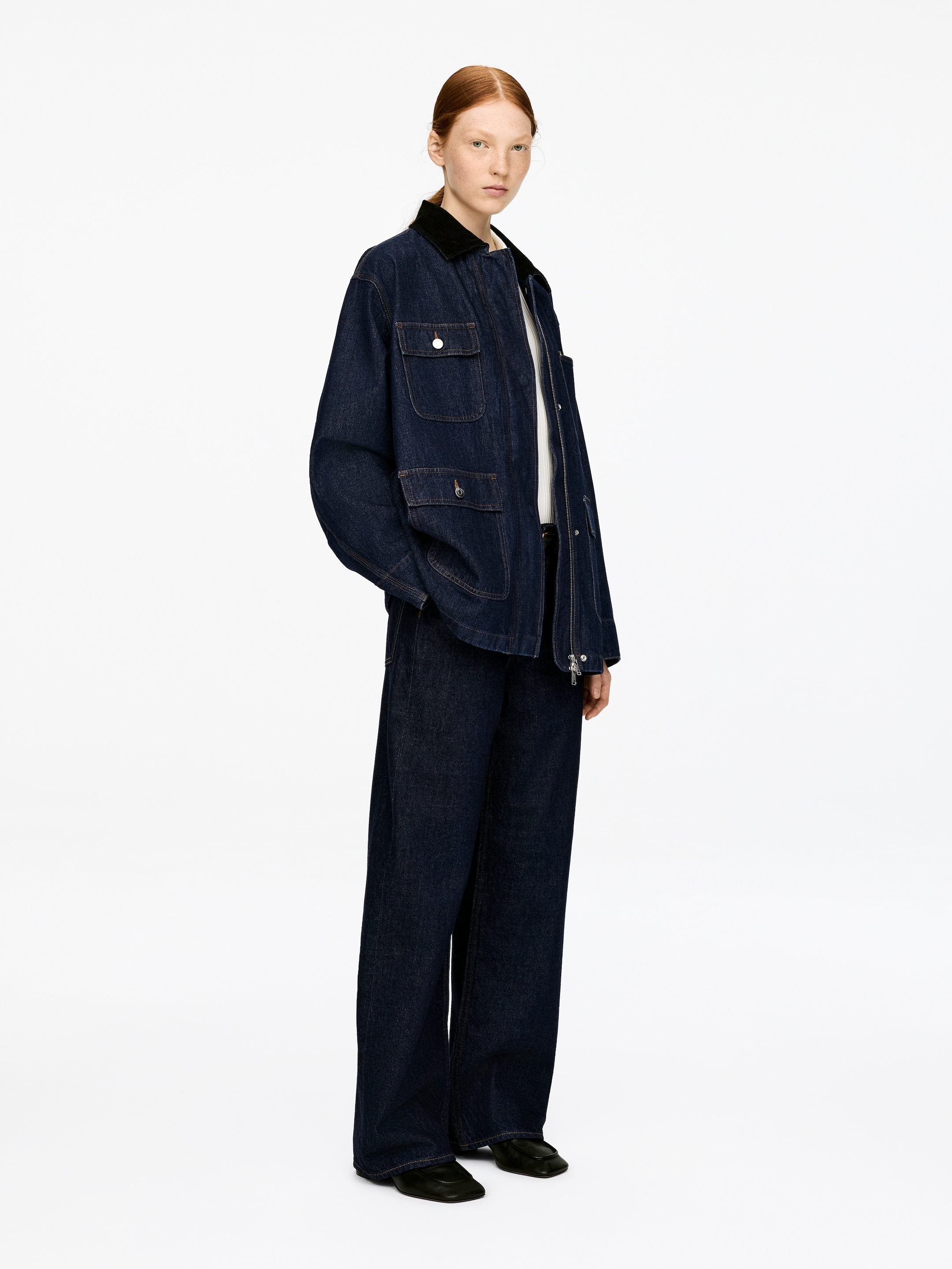 Visualizza immagine più grande: Una giacca di jeans a lavaggio scuro con un colletto in velluto a coste nero è indossata dal modello. Questa giacca oversize presenta quattro tasche a toppa sul davanti, tre delle quali con chiusura a patta e bottone, e una zip nascosta con pattina. Il modello sta mettendo una mano in una tasca a toppa inferiore.